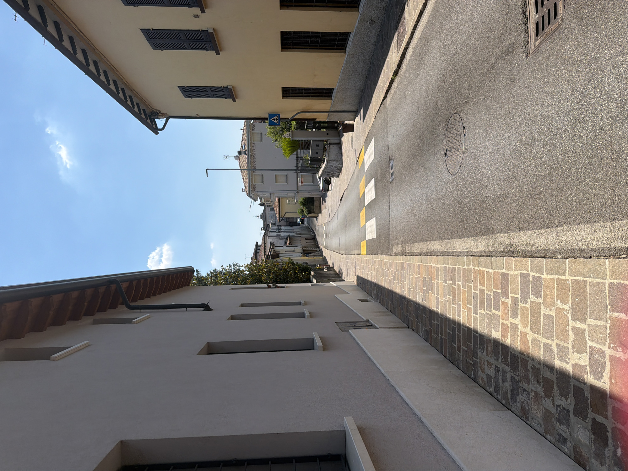 This is a street-level photograph taken on a sunny day in Padenghe sul Garda, Italy. The perspective is looking down a paved street that curves to the left, with buildings lining both sides. On the left, a light-colored building with a tiled roof overhang is prominent, its wall casting a shadow across a brick-paved sidewalk. On the right, a yellowish building with dark-shuttered windows and a similar roofline is visible.
The street itself is asphalt with a pedestrian crossing marked by yellow and white stripes. A manhole cover is present in the road. Further down the street, more buildings are visible, receding into the distance, along with trees and streetlights. A small blue and white pedestrian crossing sign is mounted on a pole near the yellowish building on the right. The sky is blue with a few scattered white clouds. The overall impression is a quiet residential or commercial street in a town.