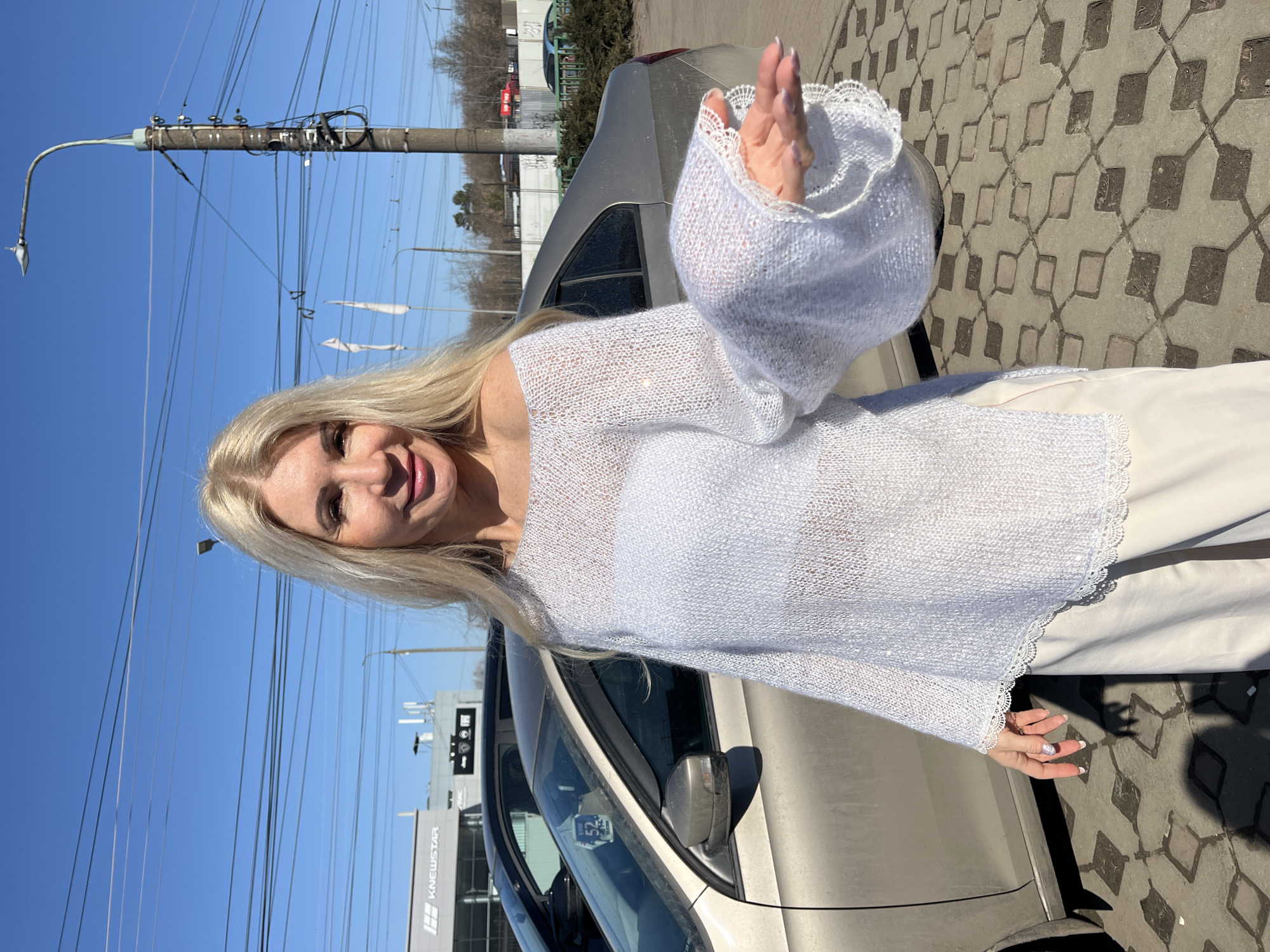 A woman is posing outdoors next to a car. She has long blonde hair and is wearing a white lace-trimmed sweater. The background includes a utility pole with wires, buildings, and a paved area with a geometric pattern. A car is parked nearby. There is a sign in the background that says "KNEWSTAR." The location is Nizhny Novgorod, Russia.