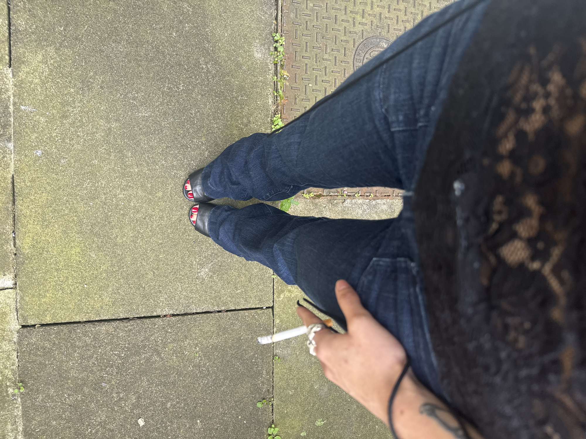 From a first-person, high-angle perspective, the image shows a person standing on a sidewalk constructed of paving stones, with some vegetation growing in the cracks. The person is wearing dark-wash jeans and black open-toe shoes with red nail polish visible. They are holding a lit cigarette in their right hand, adorned with multiple rings. A black, lace top is also visible on the person. In the upper right, a metal manhole cover is present. The location is characterized by urban elements.