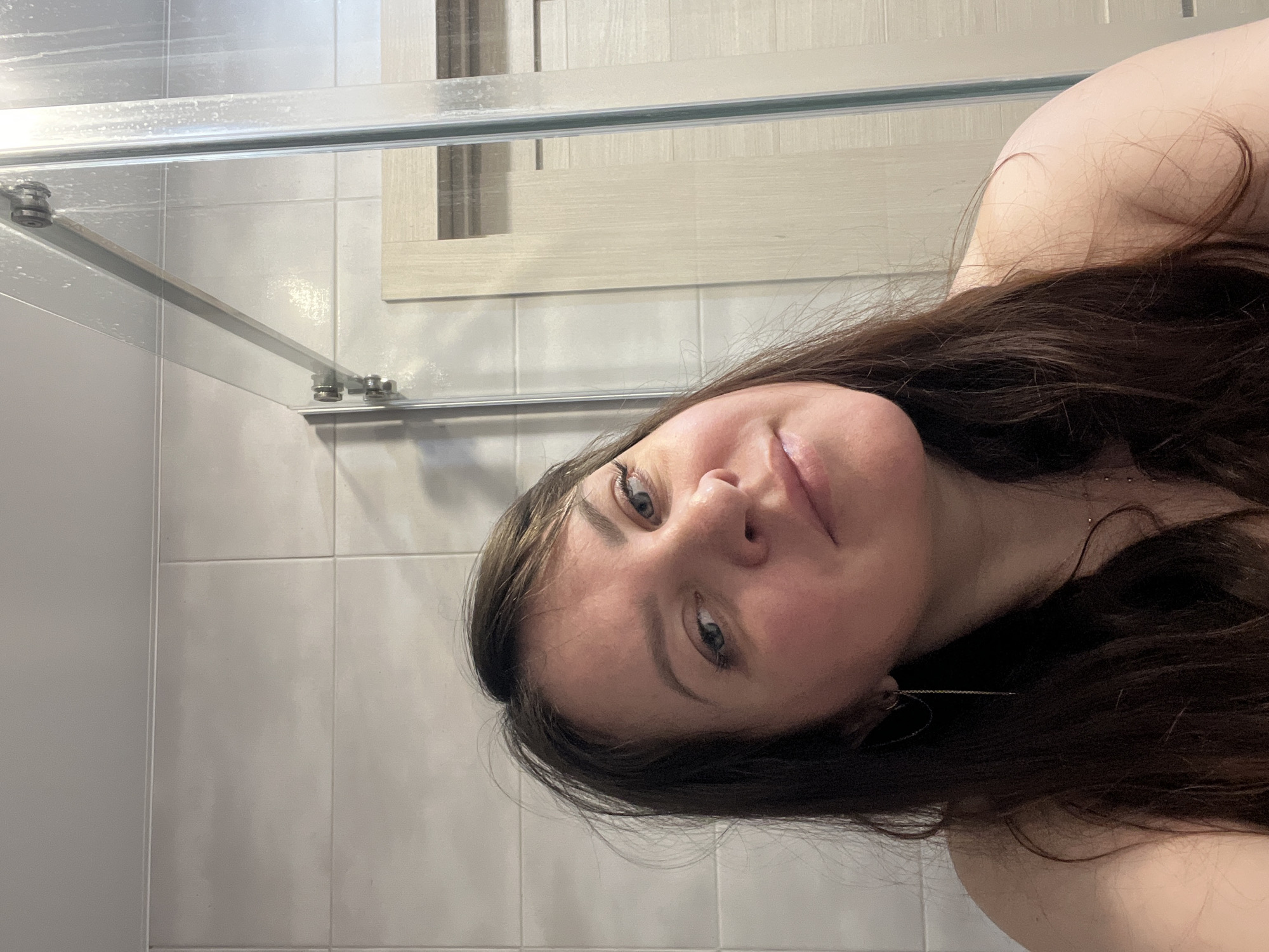 A woman with long, brown hair and fair skin is pictured in what appears to be a bathroom. The woman is angled, her face is centered in the shot with her head tilted. The background consists of light gray tiles with a shower door in the upper left of the frame. A light beige rectangular panel is affixed to the tiles, above her. The location is unspecified.