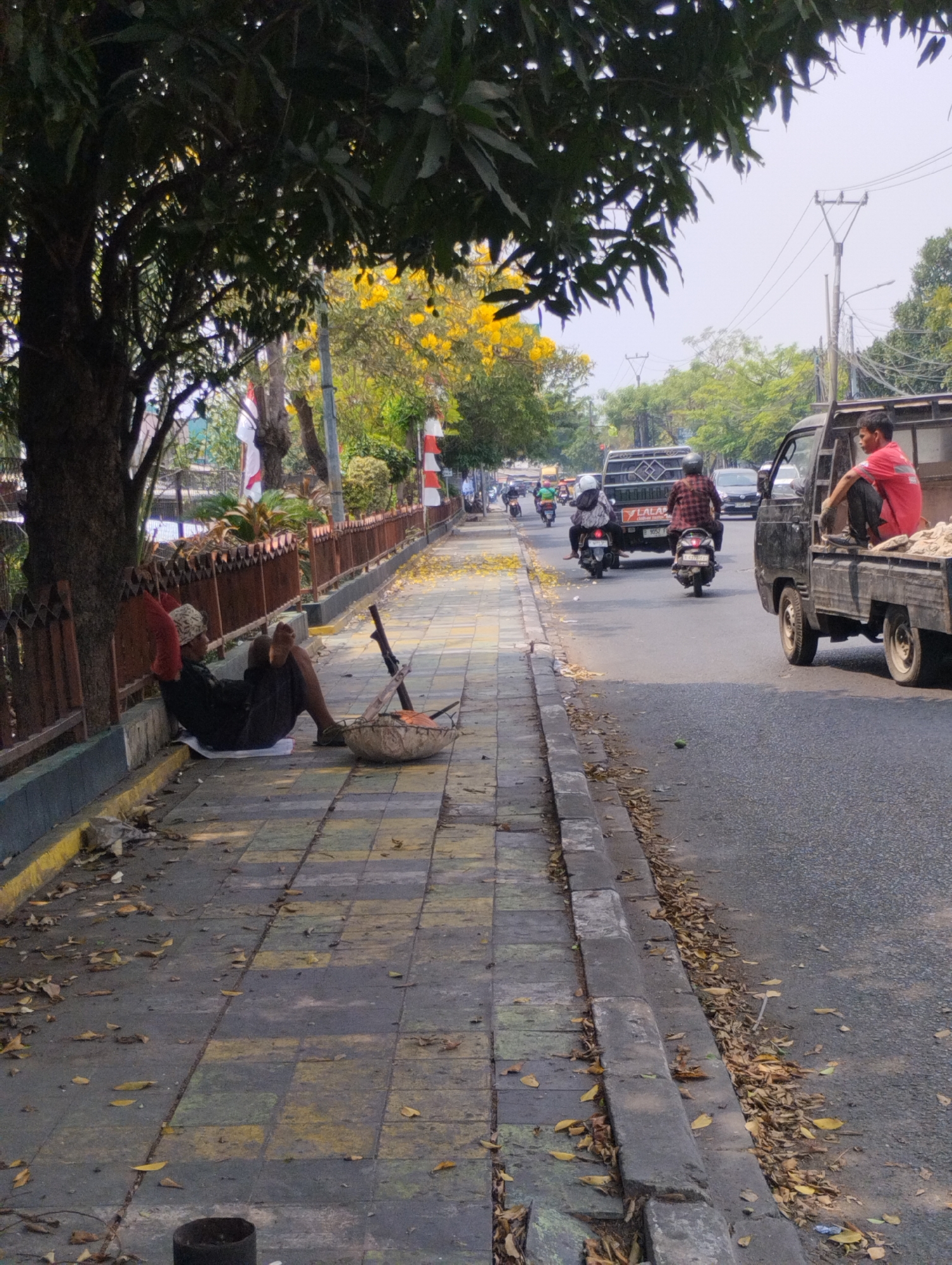 This is a ground-level, outdoor, medium-shot photograph taken on a sidewalk next to a street in Bekasi, Indonesia. A person is sitting on the sidewalk, leaning against a tree. They are wearing dark pants and a dark shirt, with a light-colored patterned head covering and a red cloth draped over their left shoulder. Their legs are extended, and they are barefoot. In front of them is a woven basket-like object with some items inside and a long stick. The sidewalk is paved with yellow and grey square tiles and is covered in scattered dry leaves. A wooden fence borders the sidewalk. The street is paved with asphalt and has a yellow line marking its edge. Several vehicles are on the street, including a black pickup truck with a person wearing a red shirt sitting in the bed, a dark-colored truck, and several motorcycles. One motorcycle has two people on it, and another has a single rider. Trees with green leaves and some with yellow flowers are visible in the background, as are utility poles and wires. The sky is overcast.