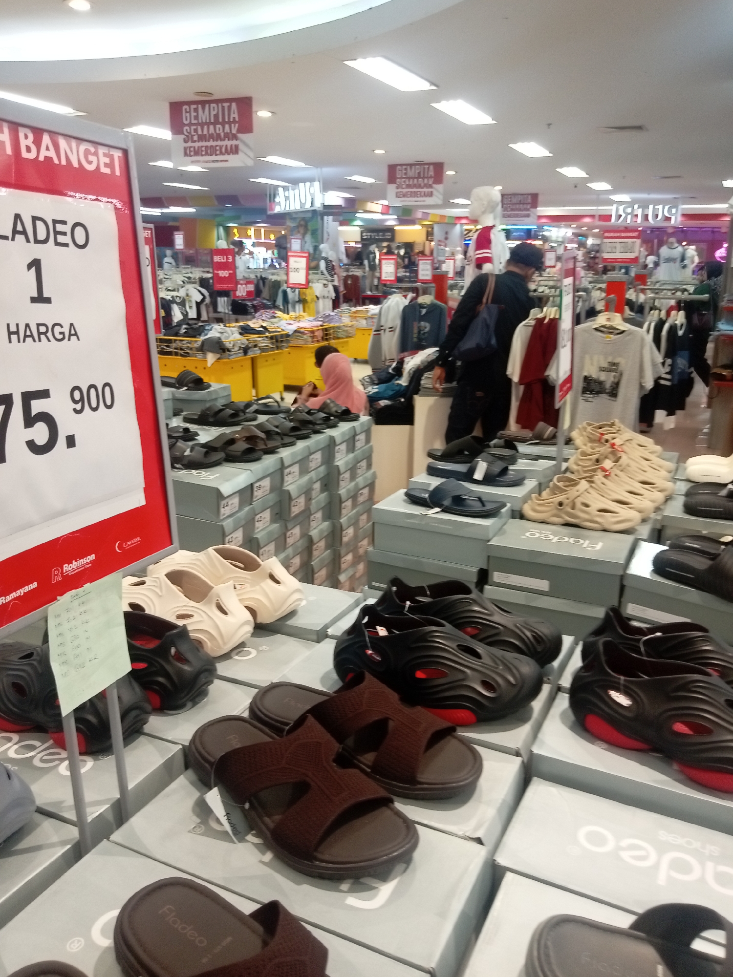 The image shows the interior of a department store in Batam, Indonesia, likely a clothing and footwear section, featuring displays of shoes on stacked boxes. In the foreground, several pairs of black and brown sandals are visible. A large red and white sign on the left advertises "LADEO 1 HARGA 75.900" (Ladeo 1 Price 75,900), indicating a sale. In the background, racks of clothing are displayed, and some shoppers are browsing. Overhead, banners with text like "GEMPITA SEMARAK KEMERDEKAAN" (Festive Independence Day) are hung, suggesting a holiday promotion. The lighting is typical of a retail environment, with overhead fluorescent lights illuminating the scene.
