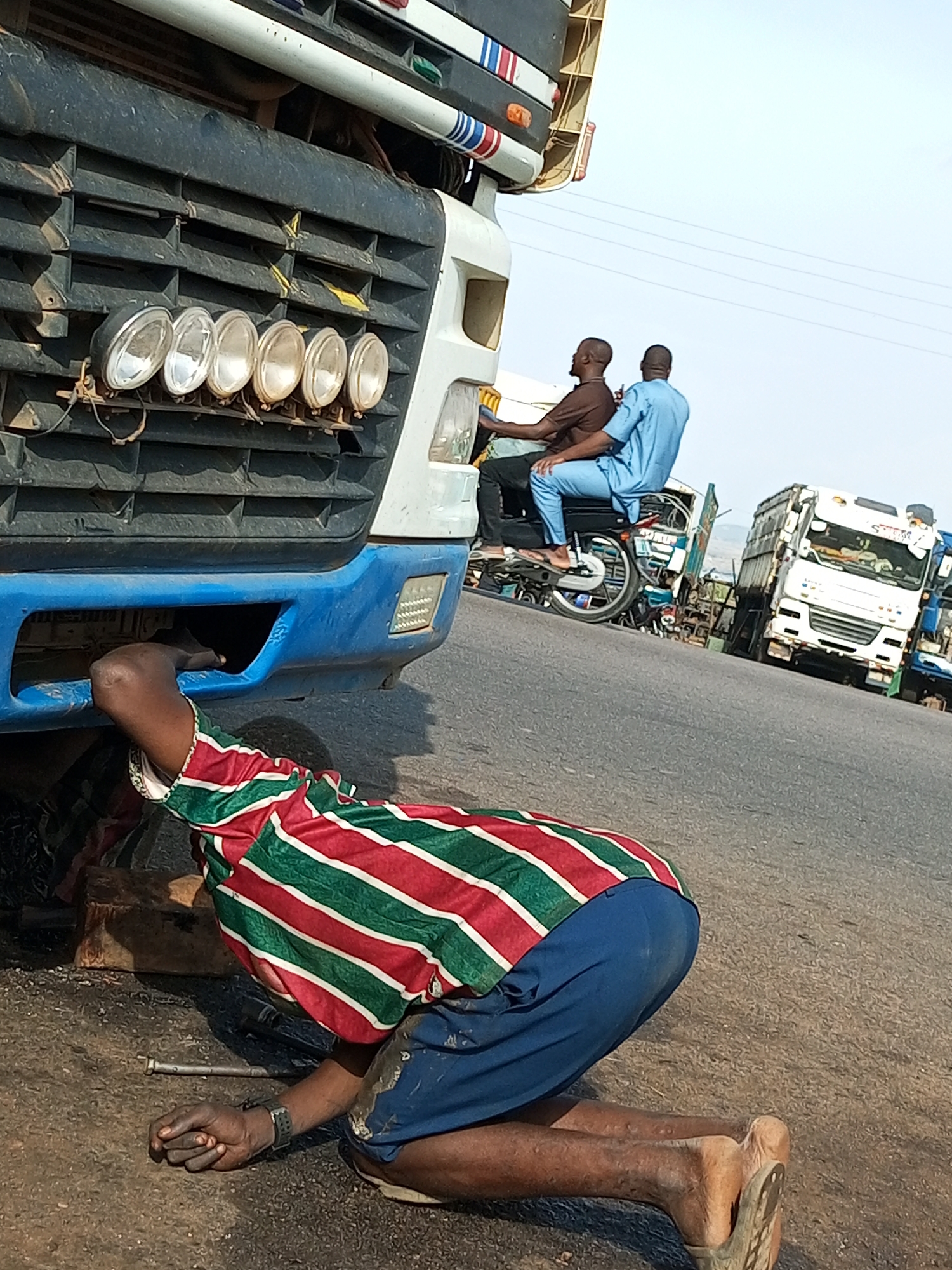 The image depicts a street scene in Gadao, Nigeria. In the foreground, a person in a striped shirt and blue trousers is kneeling, with their arm extended under a large truck for repairs. Tools, including a metal rod, are visible on the ground near their hand. In the background, two men are seated on a motorcycle. Further in the distance, other trucks are parked on the side of the road. The environment appears to be a roadside, possibly a mechanic's area, with a clear sky overhead.