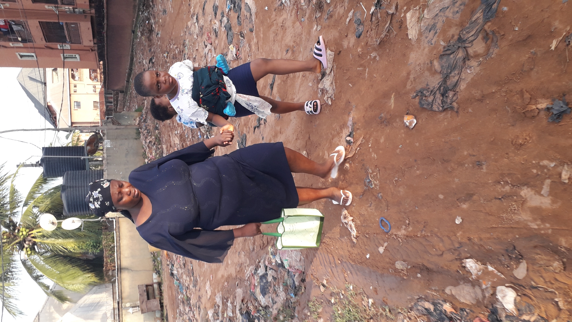 The photo depicts two people in an area littered with trash and debris in Nkpor, Nigeria. An adult woman with a dark-colored head covering and a navy blue dress is carrying a green shopping bag. A child is being held by an older child or young adolescent. The environment consists of dirt and scattered refuse. A building and a palm tree are also visible.