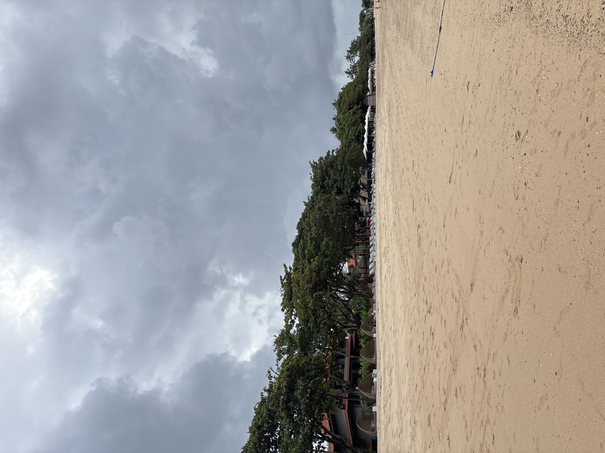 The image shows a beach in Kuta Selatan, Indonesia, under a cloudy, overcast sky. The sandy beach occupies the right side of the frame, marked with scattered debris and footprints. A row of green trees and some buildings separates the beach from the stormy sky on the left. The vegetation appears lush, indicating a tropical environment. No people are visible in the immediate scene.