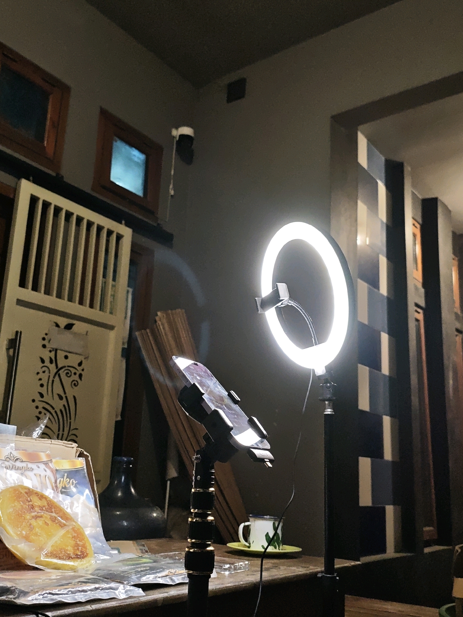 The image depicts an indoor scene, possibly a home or a small studio, with a ring light set up on a tripod. The ring light is illuminated, casting a bright light. In front of the ring light, a tablet is mounted on a flexible arm, suggesting it might be used for recording or streaming. On a wooden surface below the tablet, there is a white mug with a green cactus design, placed on a yellow saucer. To the left of the mug, there are several packaged food items, including what appears to be a flat, golden-brown pastry in clear packaging. A dark-colored bottle is visible behind the food items. In the background, a tall, slatted wooden structure, possibly a door or a screen, is present. Further to the right, a wall with a repeating pattern of dark and light rectangular tiles is visible. The overall lighting suggests it is evening or night. The location is specified as Ngasem, Indonesia.