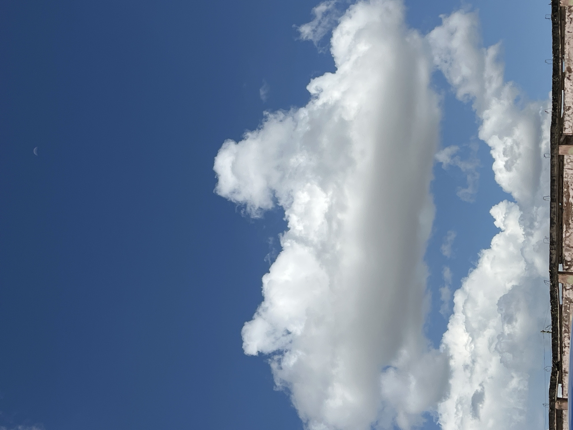 The image shows a clear blue sky with a crescent moon visible in the upper portion. A large, fluffy white cloud is centered in the middle of the frame, with smaller clouds scattered below it. The bottom of the image features the rooftop of a building with some architectural details and what appears to be construction materials or debris. The scene is not specific to Jos, Nigeria; it is a general outdoor scene.
