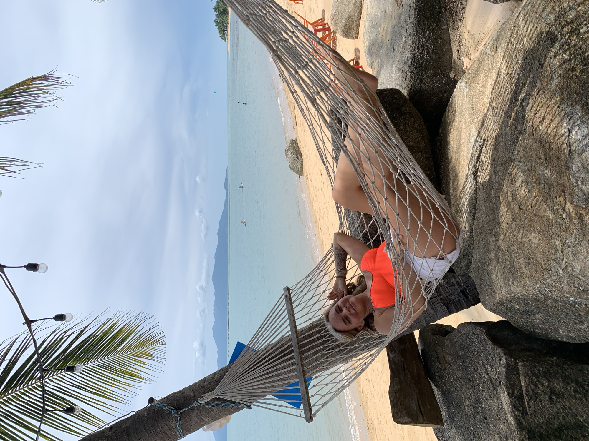 A woman is lying in a hammock tied to a palm tree on a sandy beach. She is wearing an orange tank top and white shorts, and her left arm is resting on her head. The hammock is made of netting and is supported by large rocks. In the background, the ocean stretches to the horizon, with a faint outline of mountains in the distance. The sky is overcast. The location is identified as Baan Bo Phut, Thailand.