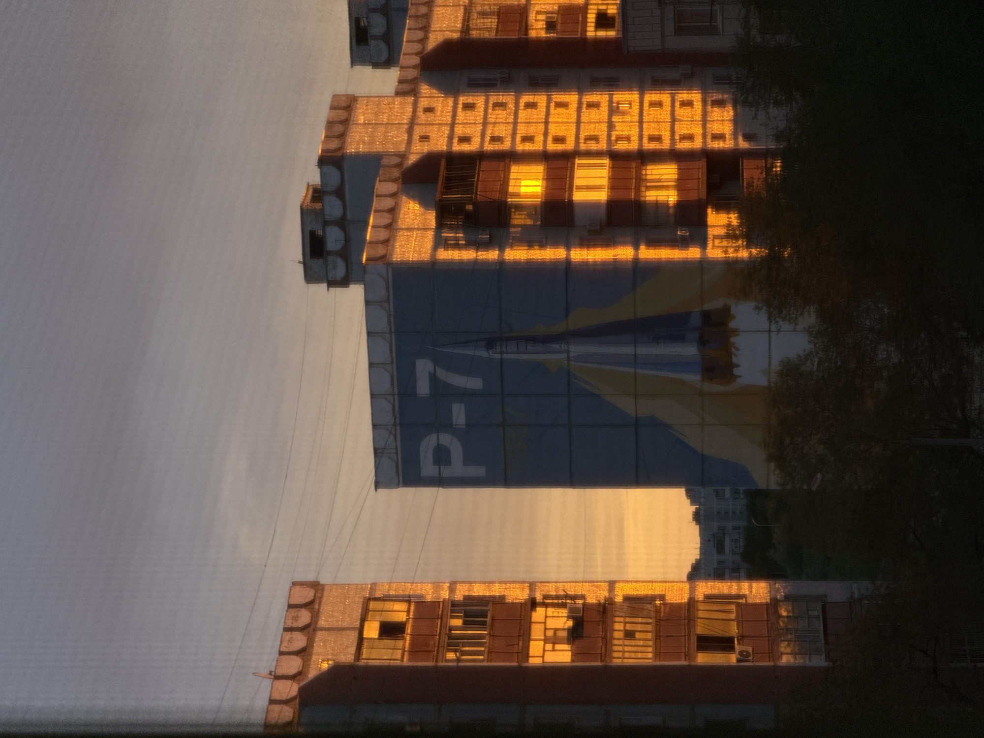 The image shows residential buildings in Baikonur, Kazakhstan, bathed in the warm glow of sunset. The sky is a gradient of soft blues and oranges. On the left, a tall apartment building with many windows reflects the golden light, with balconies visible on some floors. To the right, another apartment building is adorned with a large mural depicting a rocket, possibly referencing Baikonur's space launch history. This building also catches the sunset light, highlighting its windows and architectural details. In the foreground and between the buildings, dark silhouettes of trees obscure the lower portions of the structures. The overall scene is tranquil and captures a moment of urban beauty during twilight.