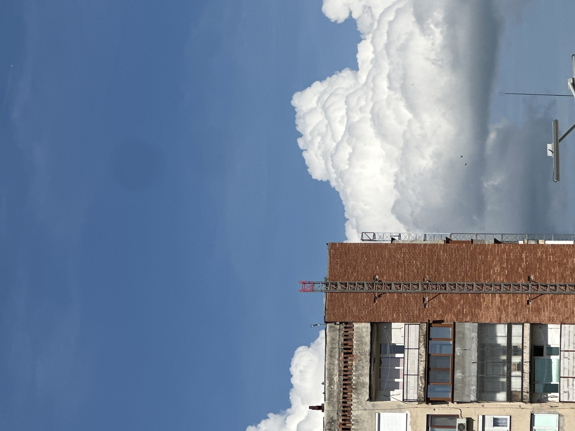 The image displays the upper section of a building against a vivid sky, likely in Lovech, Bulgaria. The lower portion of the frame features the building's facade, characterized by a prominent brown, tiled section at its top, beneath a flat roof. A metallic truss structure, possibly part of a small crane or scaffolding, extends horizontally from the building's top edge, with a distinct red element at its left end. Below the tiled section, the facade consists of concrete or stucco with multiple windows and enclosed balconies, exhibiting varying designs and reflections. An air conditioning unit is visible on a lower level of the building.
The upper part of the image is dominated by a bright blue sky, which is particularly clear on the left. Large, voluminous white cumulus clouds occupy the right side, creating a striking contrast with the blue. These clouds appear to be building up, with some darker areas hinting at their depth or partial shadowing. A small white speck, possibly a distant plane or bird, is faintly visible in the clear blue expanse on the far left. Two smaller, dark, bird-like shapes are seen flying closer to the large cloud formation. A utility pole or antenna structure extends into the sky from the far right edge of the frame.
The scene depicts a bright, sunny day, likely mid-day, indicated by the strong illumination and the nature of the clouds. No people or specific activities are visible, and no text is discernible.
