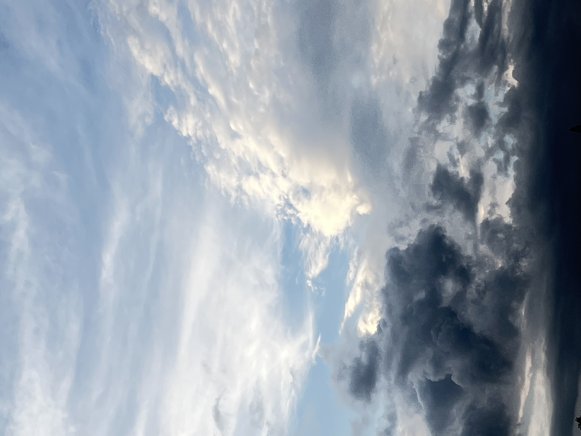 The image displays a dramatic sky over what appears to be a landscape or cityscape, with dark, ominous clouds dominating the lower portion and lighter, wispy clouds filling the upper sky. The contrast suggests a change in weather, possibly an approaching storm. There are no discernible people or specific objects that would definitively place the location in Krasnodar, Russia. The scene is solely focused on the atmospheric conditions.