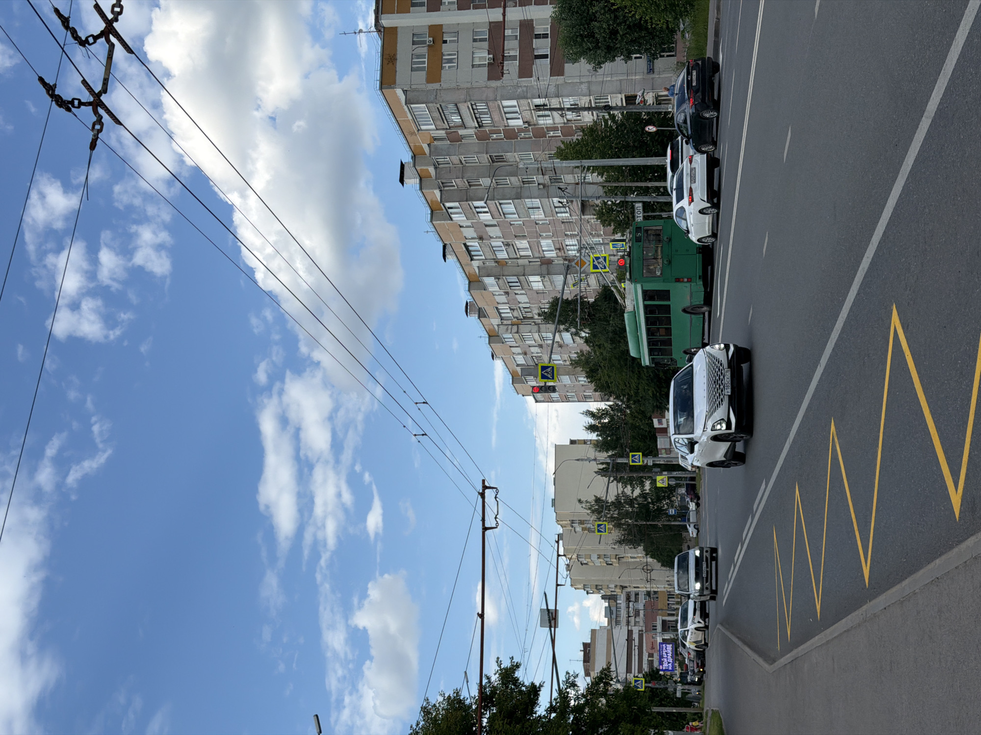 A street scene depicts a blue sky with scattered white clouds. Overhead, a network of trolley wires crisscrosses the sky. In the foreground, a road curves to the left with yellow zigzag markings on the asphalt. Several cars are visible on the road, including a white SUV in the right lane and a white sedan in the left lane. Further down the road, a green trolleybus is stopped at an intersection. On the right side of the road, a tall, multi-story apartment building with a grey facade and yellow trim dominates the background. Trees line the sidewalk in front of the building. Pedestrian crossing signs with yellow diamond shapes are visible at the intersection. The overall impression is of a typical urban street on a cloudy day.