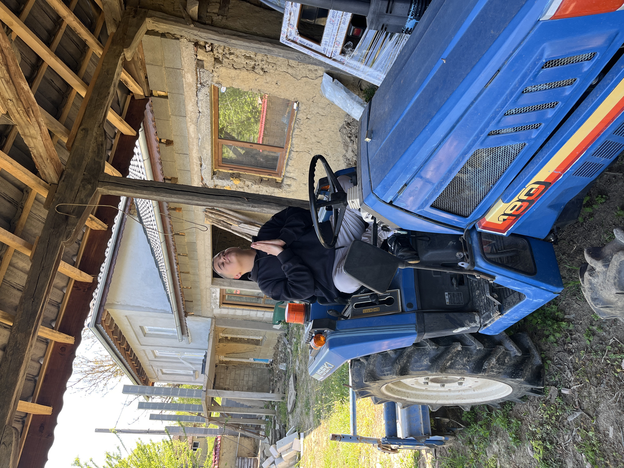 A person is sitting on a blue Iseki tractor, possibly a model 190, in an outdoor setting. The person is wearing a dark hooded sweatshirt and light-colored pants, with their hands together. The surrounding environment includes a dilapidated building with a wooden-framed window and a partially collapsed roof structure. Newer building materials are present, suggesting ongoing construction or renovation. The location appears to be a rural area, possibly in the vicinity of Rish, Bulgaria, based on the architectural style of the buildings. The ground is covered with grass and dirt.