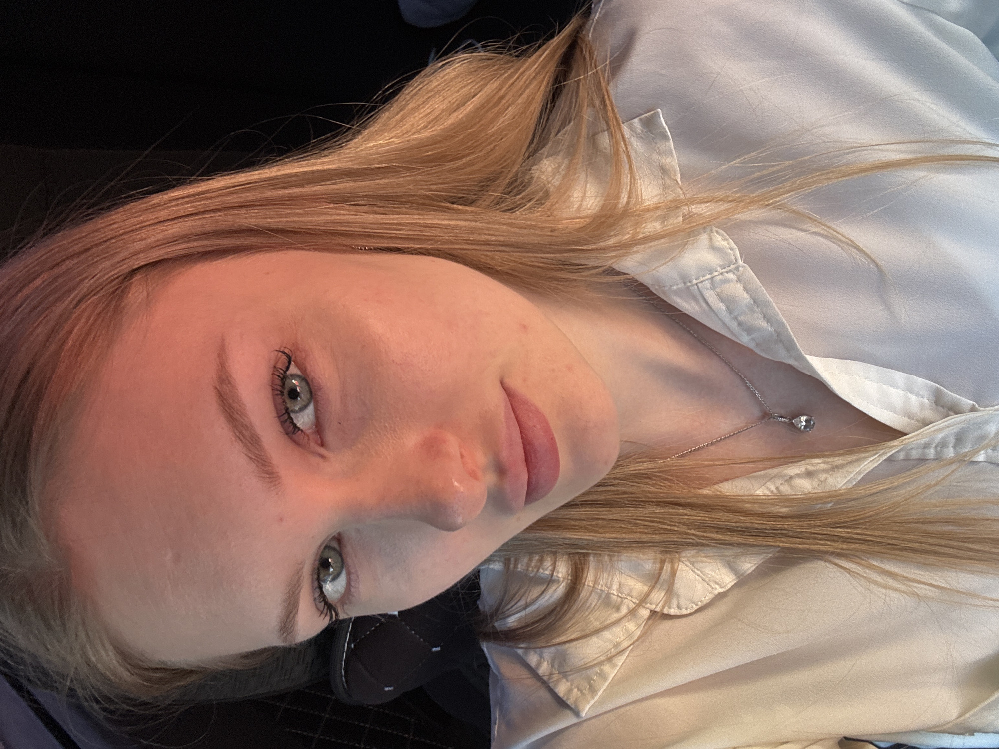 A blonde-haired young woman with light skin and grey eyes is shown in a close-up, tilted, indoor shot. She wears a white button-down shirt and a silver necklace with a small pendant. A black surface, possibly car seat upholstery, is visible in the lower left corner. No information about the location is discernible from the image.
