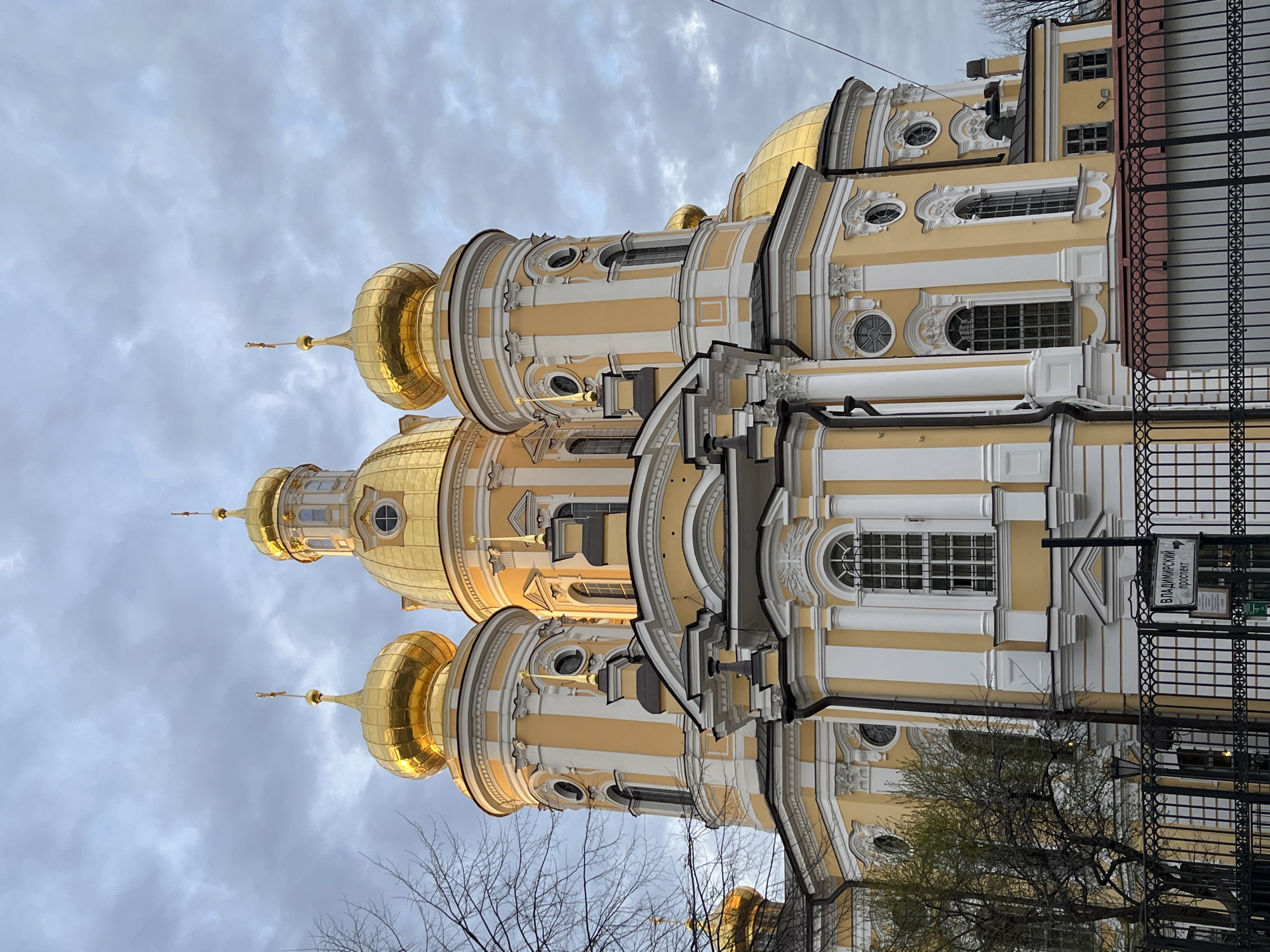 The image captures a close-up, slightly angled view of the Saint Nicholas Naval Cathedral in St. Petersburg, Russia. The cathedral's architectural elements are prominently displayed, showcasing its golden domes, intricate stucco work, and cream-colored walls with yellow accents. The domes glisten, reflecting the ambient light of the overcast sky. The architectural style is Baroque. The foreground includes bare tree branches. A wrought iron fence with a sign is visible on the right side of the frame. The sign has Russian text. The overall impression is that of a grand, ornate structure set against a cloudy backdrop.
