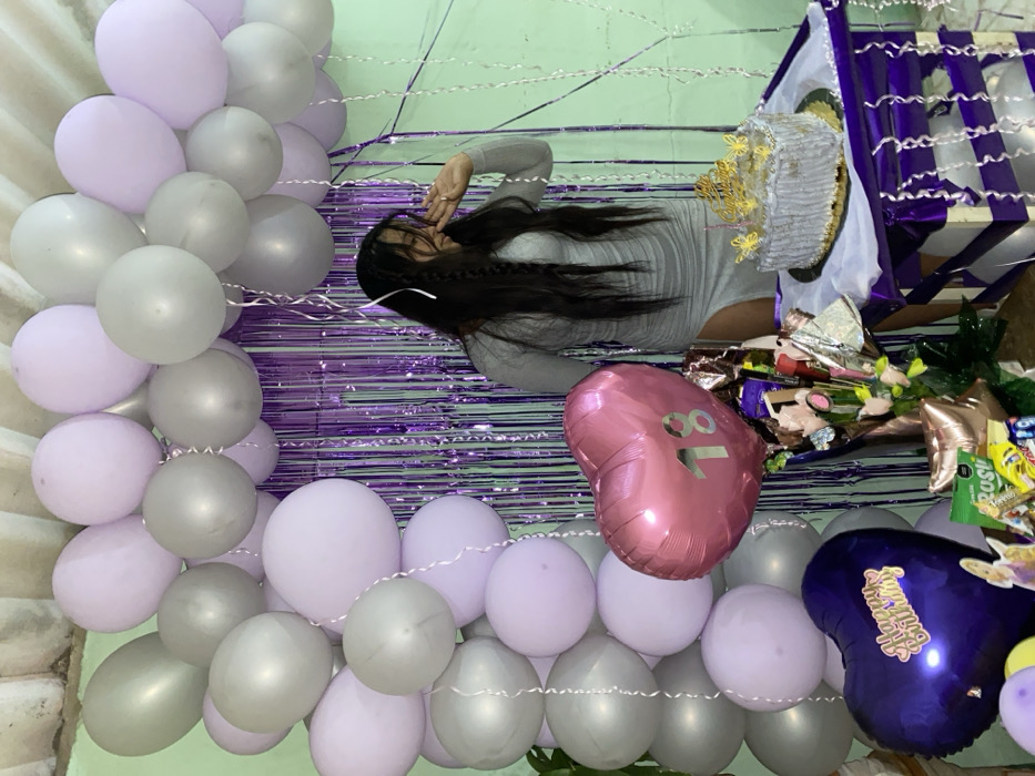 A young woman poses with a birthday celebration setup. She's wearing a gray dress and dark hair is braided. Purple tinsel hangs as a backdrop. A balloon arch in shades of lavender and gray frames the scene. Gifts, including a wrapped cake, chocolates, and flowers, are displayed. A pink heart-shaped balloon with "18" printed on it and a dark purple balloon with "Happy Birthday" add to the festive atmosphere.