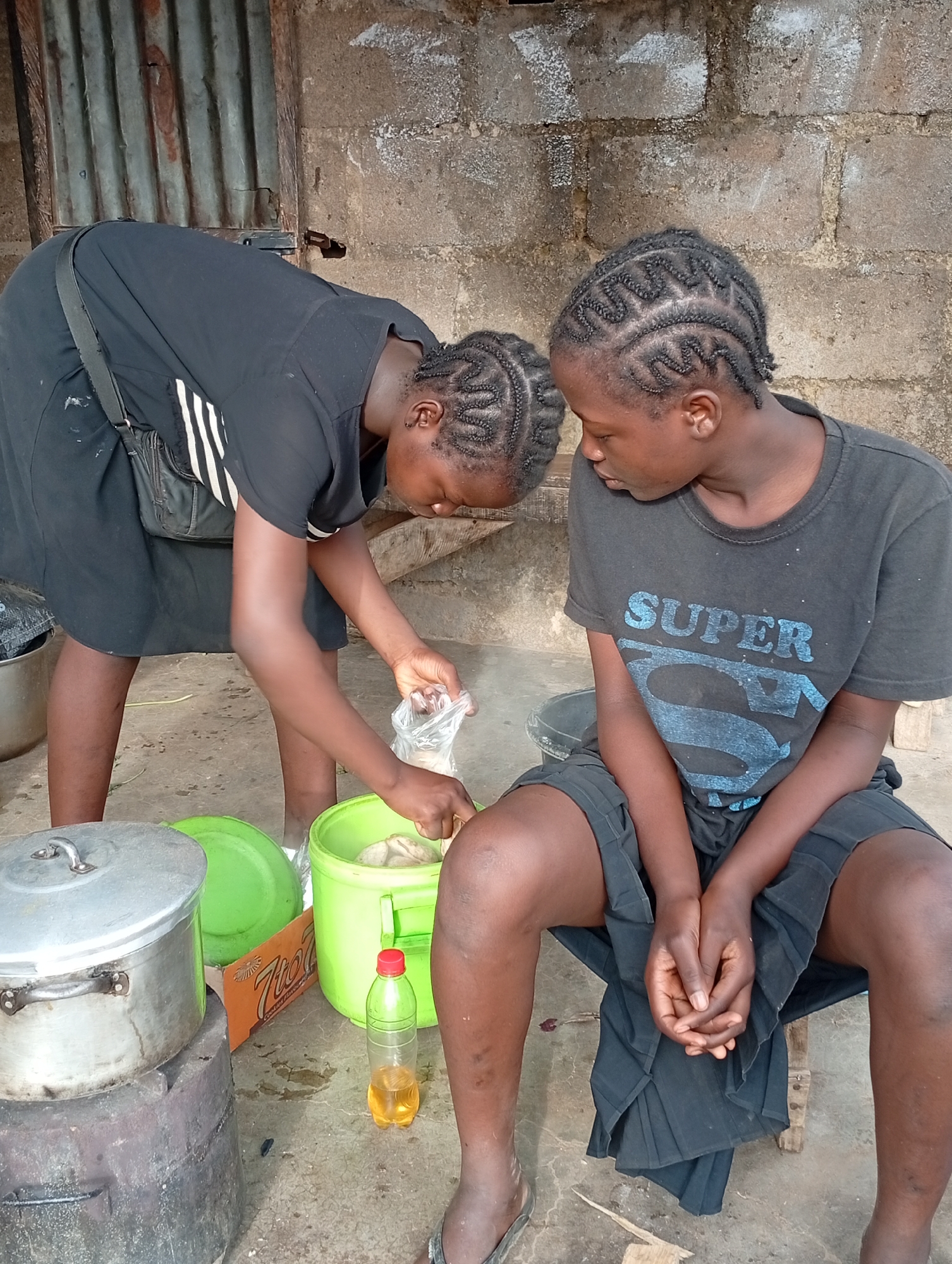 Two young women are present in the image. One is bending over a green plastic bucket, holding a plastic bag, seemingly placing contents from the bag into the bucket. She is wearing a black dress with white stripes on the sleeves and a shoulder bag. The other woman is seated on a wooden stool, wearing a dark-colored t-shirt with the word "SUPER" on it and dark shorts. Her hands are clasped together in her lap. To the left of the image is a stack of cooking pots on a charcoal stove, next to a green lid, an open cardboard box, and a plastic bottle with yellow liquid. The setting appears to be outdoors, with a concrete floor and a wall made of brick or concrete blocks in the background. The city and location are unknown.