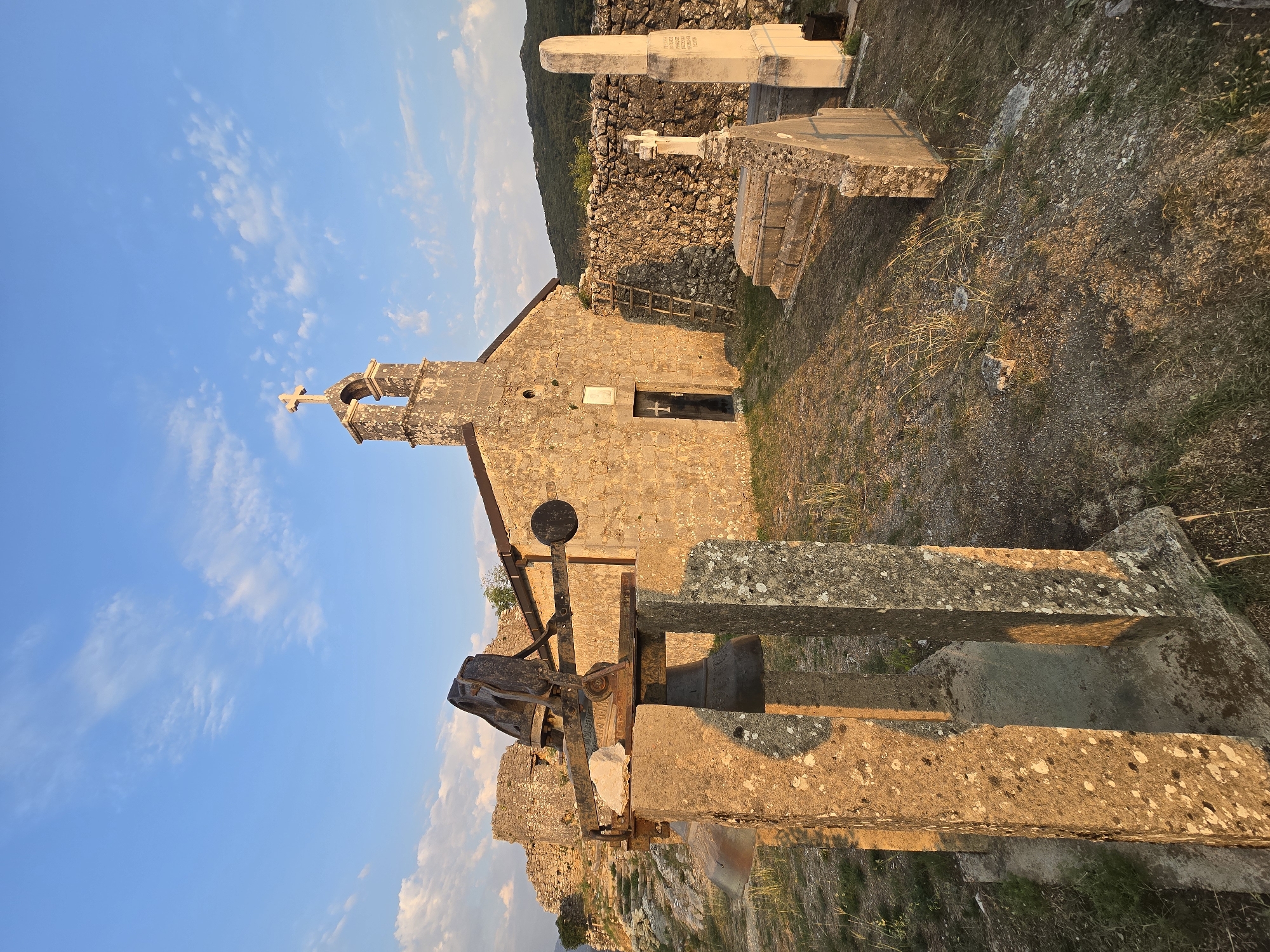 In the village of Gornja Vrbica, Montenegro, a stone church stands atop a hill against a backdrop of a clear blue sky with scattered white clouds. The church has a simple facade with a single wooden door and a bell tower adorned with a cross. In the foreground, a weathered bell rests on a stone pedestal, illuminated by the warm glow of the setting sun. Surrounding the church are remnants of stone walls and several gravestones, suggesting a small cemetery. The landscape is rugged, with sparse vegetation and distant hills. The scene conveys a sense of timelessness and tranquility.