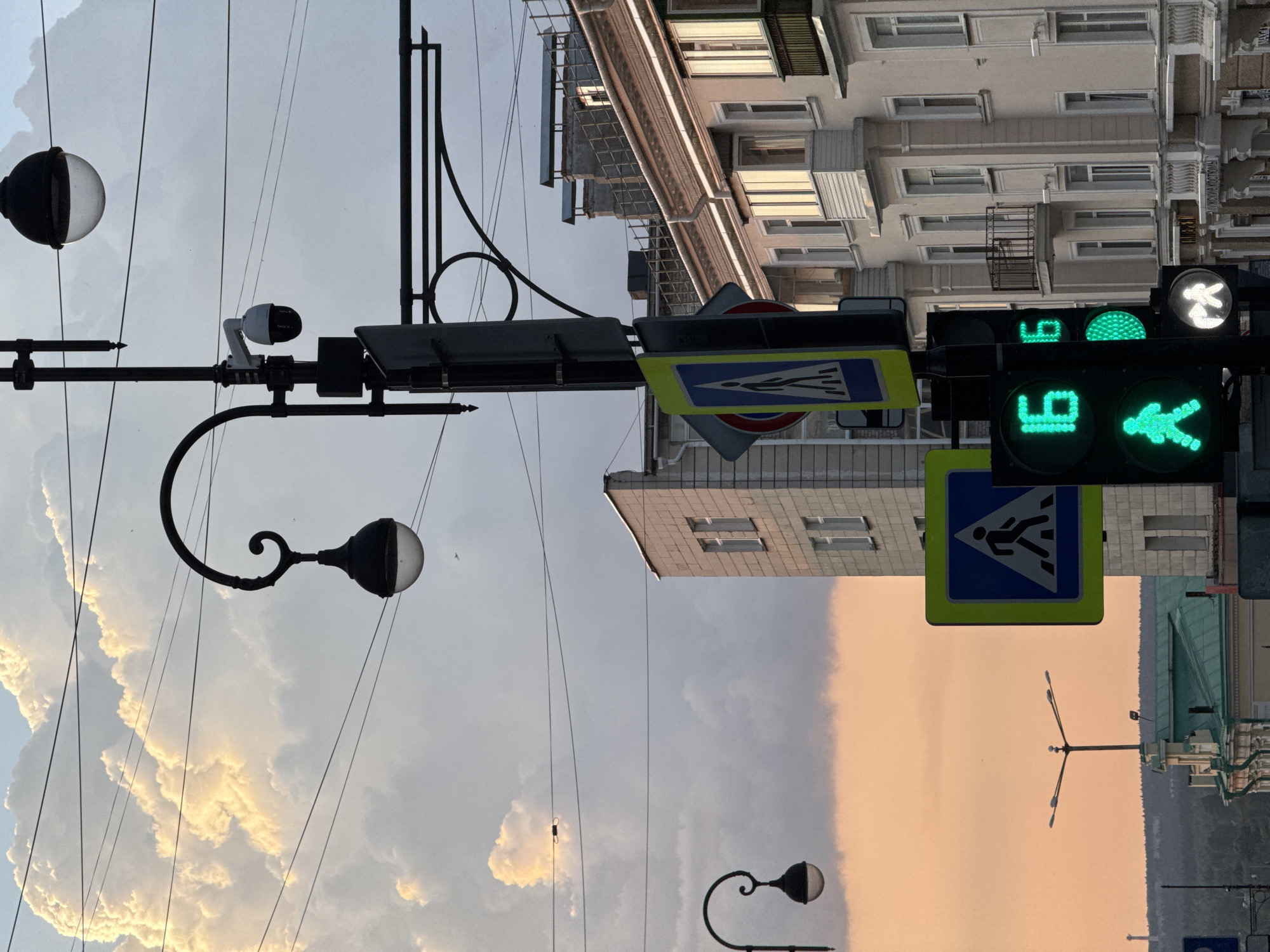 The image depicts a street scene in Perm, Russia, with a focus on a traffic light pole and pedestrian crossing signals. The traffic light displays green for both vehicles and pedestrians, indicated by illuminated figures of walking people and the numbers "16" and "6." A pedestrian crossing sign, showing a person walking, is visible on the pole. The background features an urban setting with buildings, street lamps, and a sky filled with large, dramatic clouds tinged with the orange and yellow hues of sunset. Overhead wires crisscross the sky. One building in the background has a light-colored facade with balconies and windows. Another, closer building, also appears to be made of light-colored bricks. The overall impression is of a city street at dusk, with the traffic signals and pedestrian crossings prominently featured.