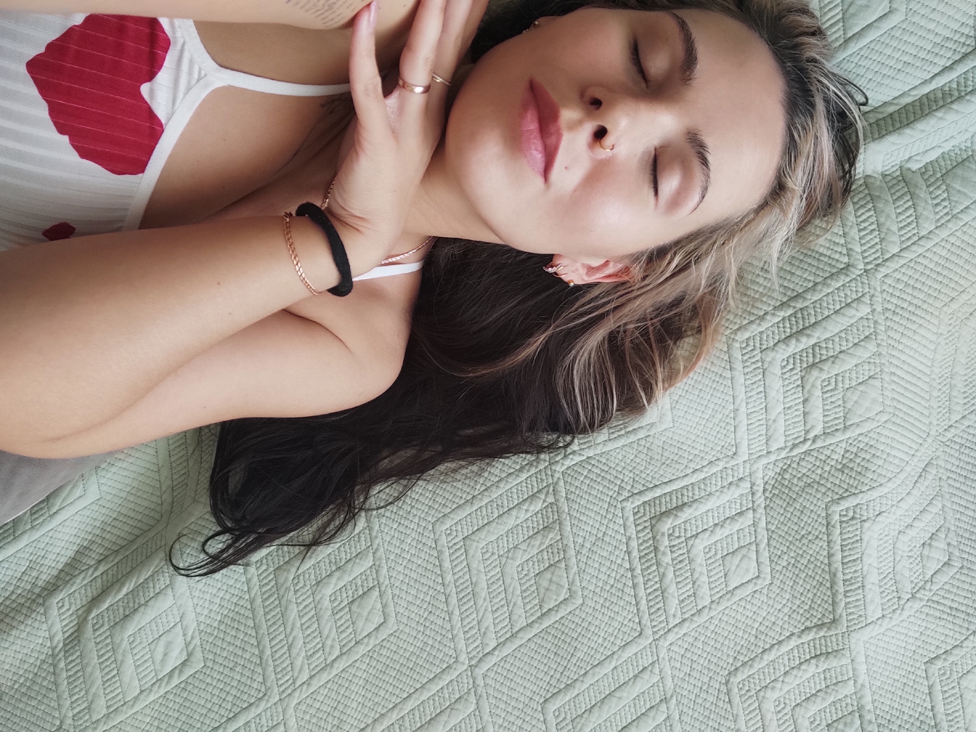 A woman with dark and blonde hair lies on a light green quilted bedspread. She wears a white tank top with a red heart design. Her eyes are closed and her hand is near her face with rings on her fingers. She has a nose ring and earrings. She is wearing a gold bracelet and a black hair tie on her other wrist. The image appears to be taken from an overhead perspective. There is no specific evidence to suggest the image was taken in Chaikovsky, Russia.