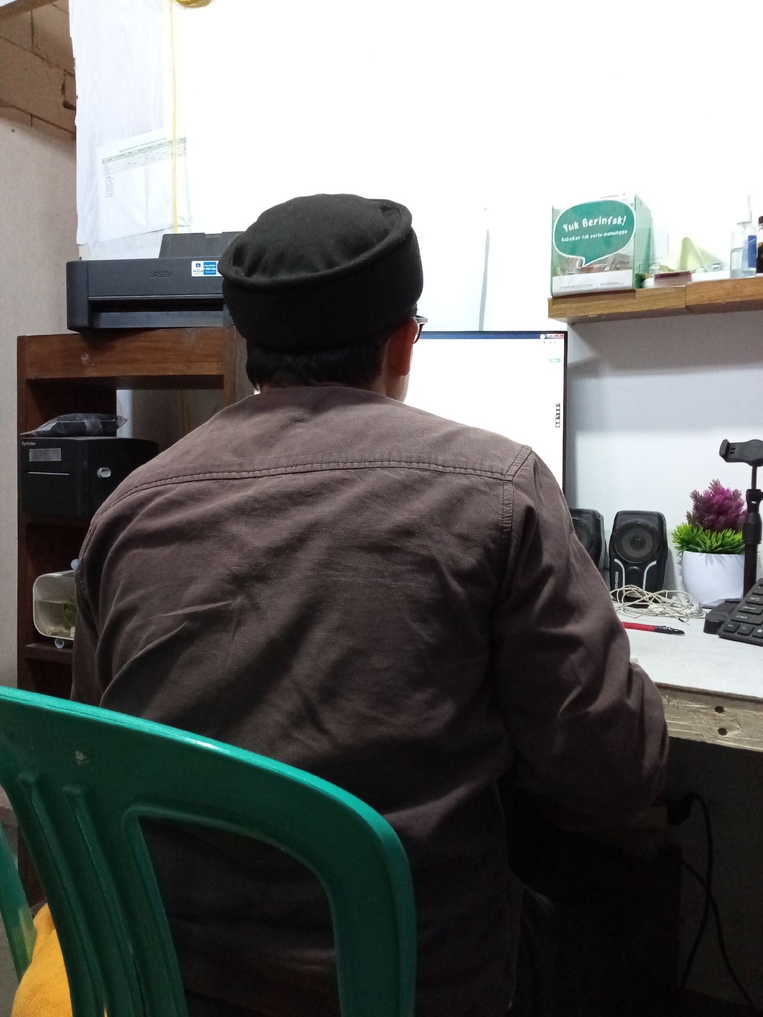 A person wearing a black cap and a brown long-sleeved shirt is seated at a desk in front of a computer monitor. The person is viewed from behind. On the desk are two speakers, a potted plant, a keyboard, a pen, and a phone. To the left of the desk, on a wooden shelf, is a printer and a small aquarium. The room appears to be an office or study. This scene is located in Cihanjuang, Indonesia.