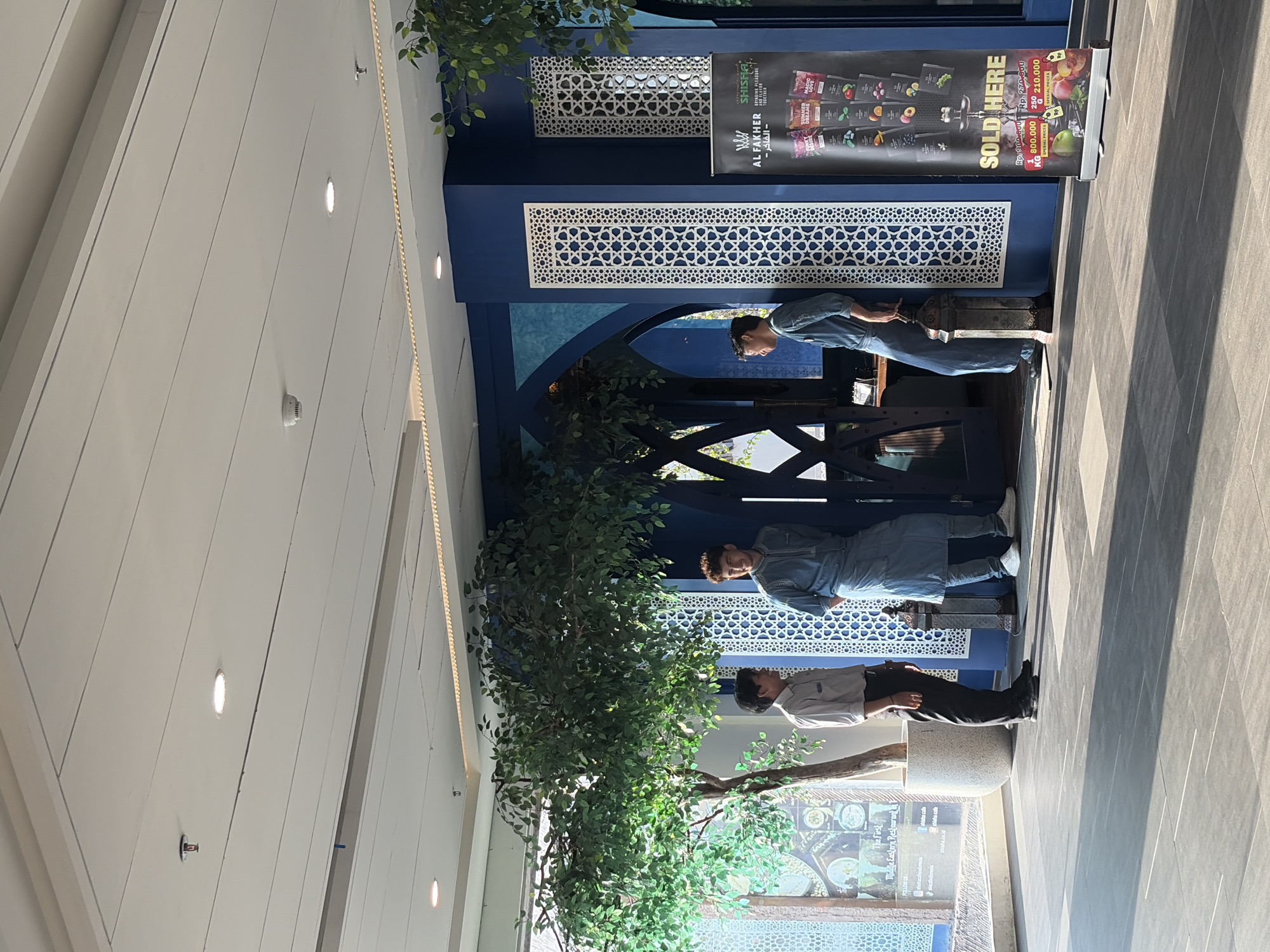 Three men are standing in an indoor environment. One man on the left is wearing a light-colored collared shirt and dark pants. The other two men are wearing similar light blue traditional garments. They appear to be conversing. In the background, there are decorative blue walls with white patterned panels, and large artificial trees. A promotional banner for "Al Fakher Shisha" is visible on the right. The scene suggests a retail or restaurant setting, likely in Kuta, Indonesia, given the cultural attire and banner. The lighting suggests it is daytime.