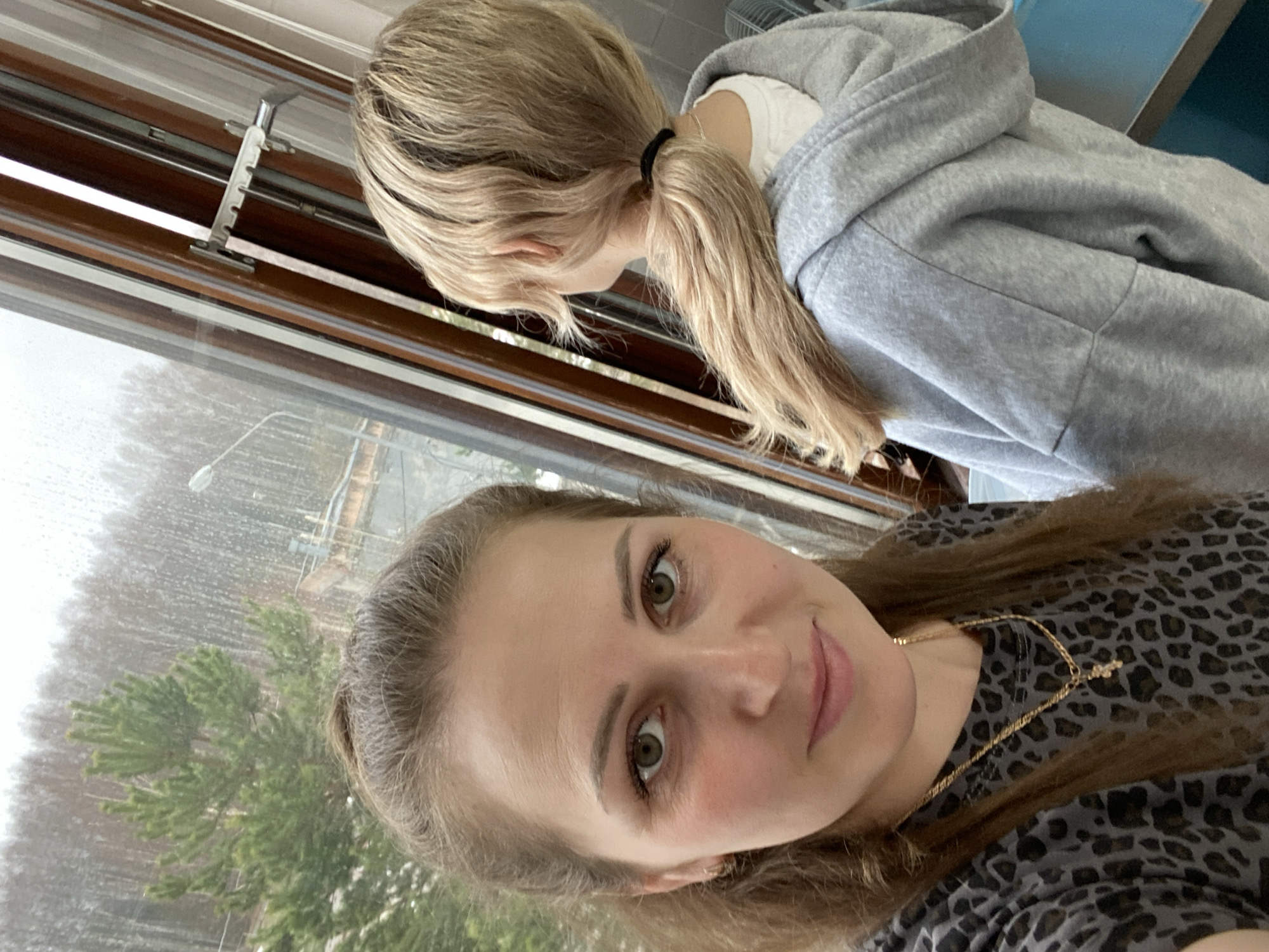 A selfie shows two women in Марковский, Russia. The woman in the foreground has brown hair and is wearing a leopard-print shirt and a gold necklace. Her face is turned slightly to the side, and she is smiling. Behind her is a window with a view of a rainy landscape with trees. The window frame is brown. To the right of the window, another woman with blonde hair in a ponytail is partially visible. She is wearing a gray hoodie and a white shirt. The background shows the city in the rain, with trees visible through the glass.