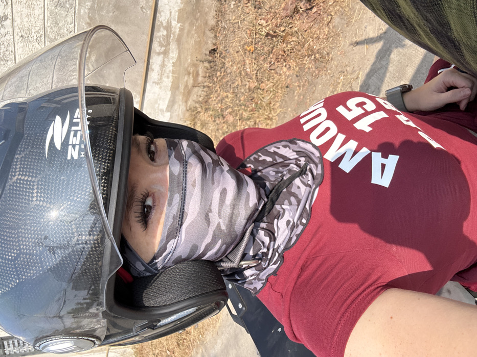 A person wearing a black motorcycle helmet with a clear visor and a camouflage face mask is pictured outdoors. They wear a maroon shirt with white text reading "FAMOUS 15". A smartwatch is visible on their left wrist. The background includes pavement and dry, brown grass or weeds. The photo is taken from a high angle, angled to the side.
