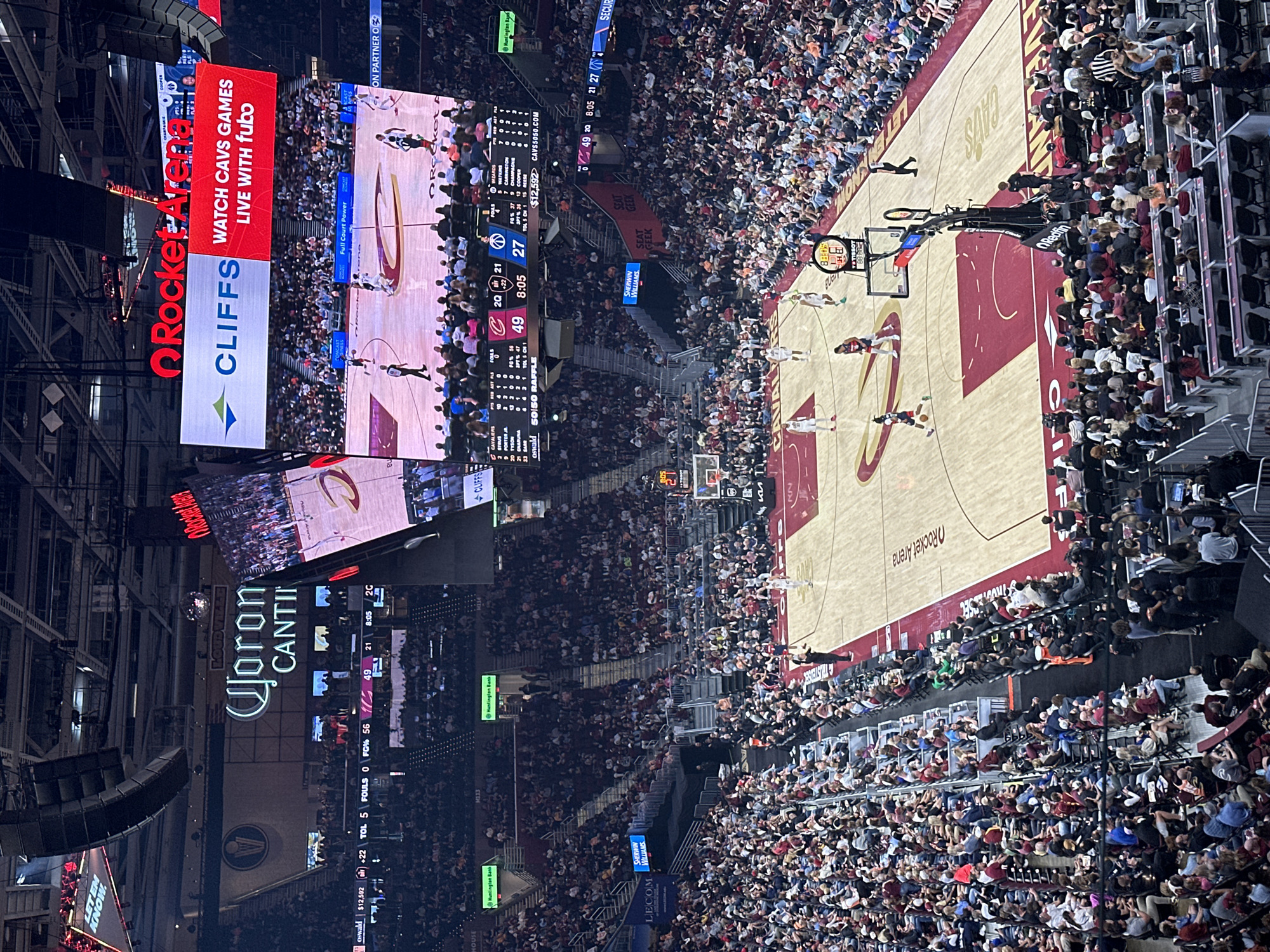 The image captures a basketball game in progress at Rocket Mortgage FieldHouse, Cleveland, Ohio, United States. The arena is packed with spectators, mostly in casual attire. The basketball court is visible from an elevated angle, including players, referees, and team logos. Overhead, large digital screens display live footage, game statistics, and advertisements from sponsors such as Rocket Mortgage, Cliffs, Corona Cantina, Sherwin Williams, Huntington Bank, SeatGeek and Lecom. The score is displayed as Cavaliers 49 and the opposing team is at 27, with 8:05 remaining in the 2nd quarter.