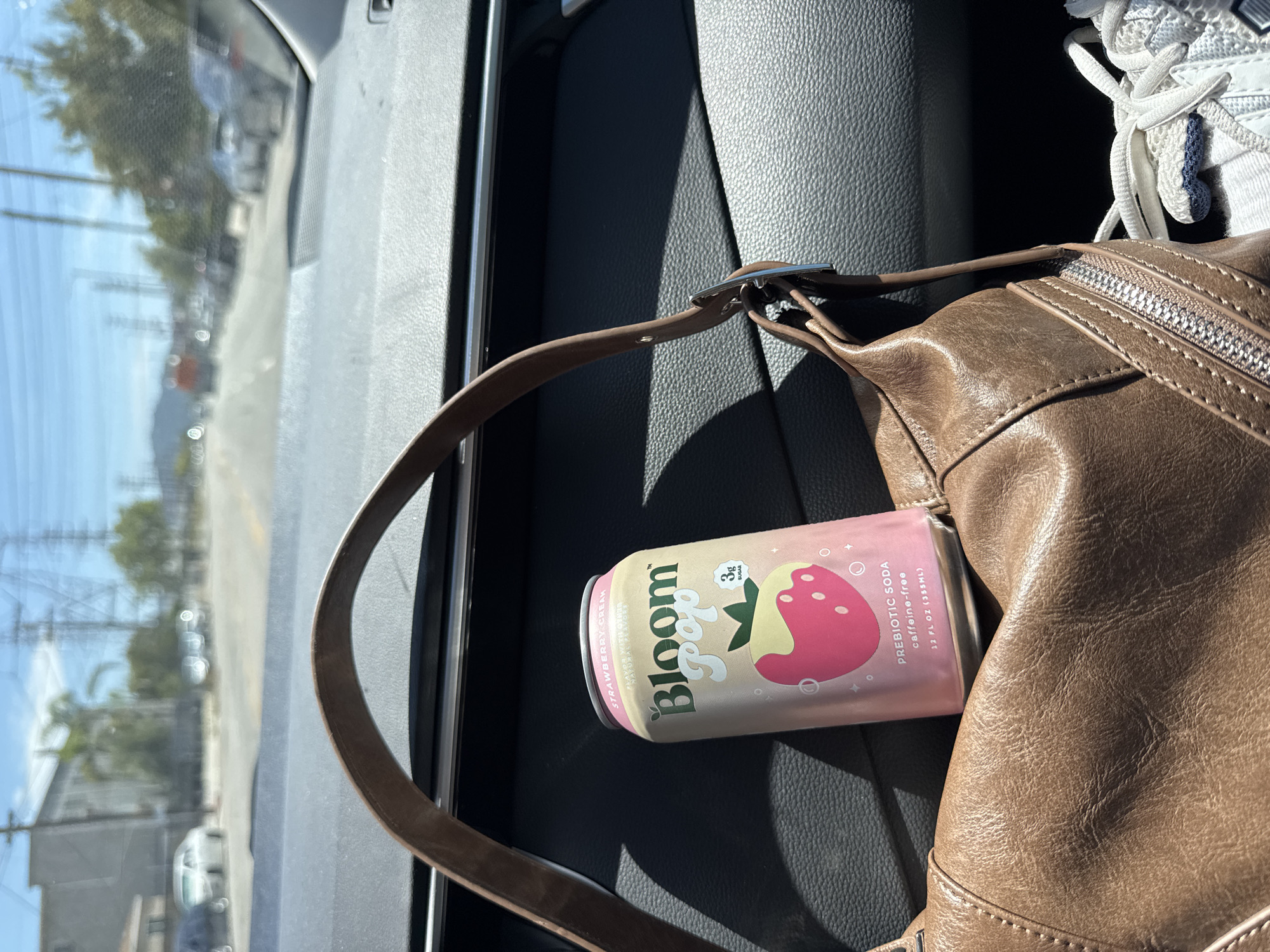 Here is a description of the image:
The image shows the inside of a car with a view of a street scene outside the window. A can of "Bloom pop" prebiotic soda (Strawberry Cream flavor) is resting on the car seat between a brown leather handbag and what appears to be a white sneaker. The view out the window suggests an urban area, possibly in Los Angeles, United States, given the style of buildings, palm trees, and utility poles. There are cars parked along the street visible through the window. The photo is taken in daylight, with the sunlight illuminating parts of the car's interior.