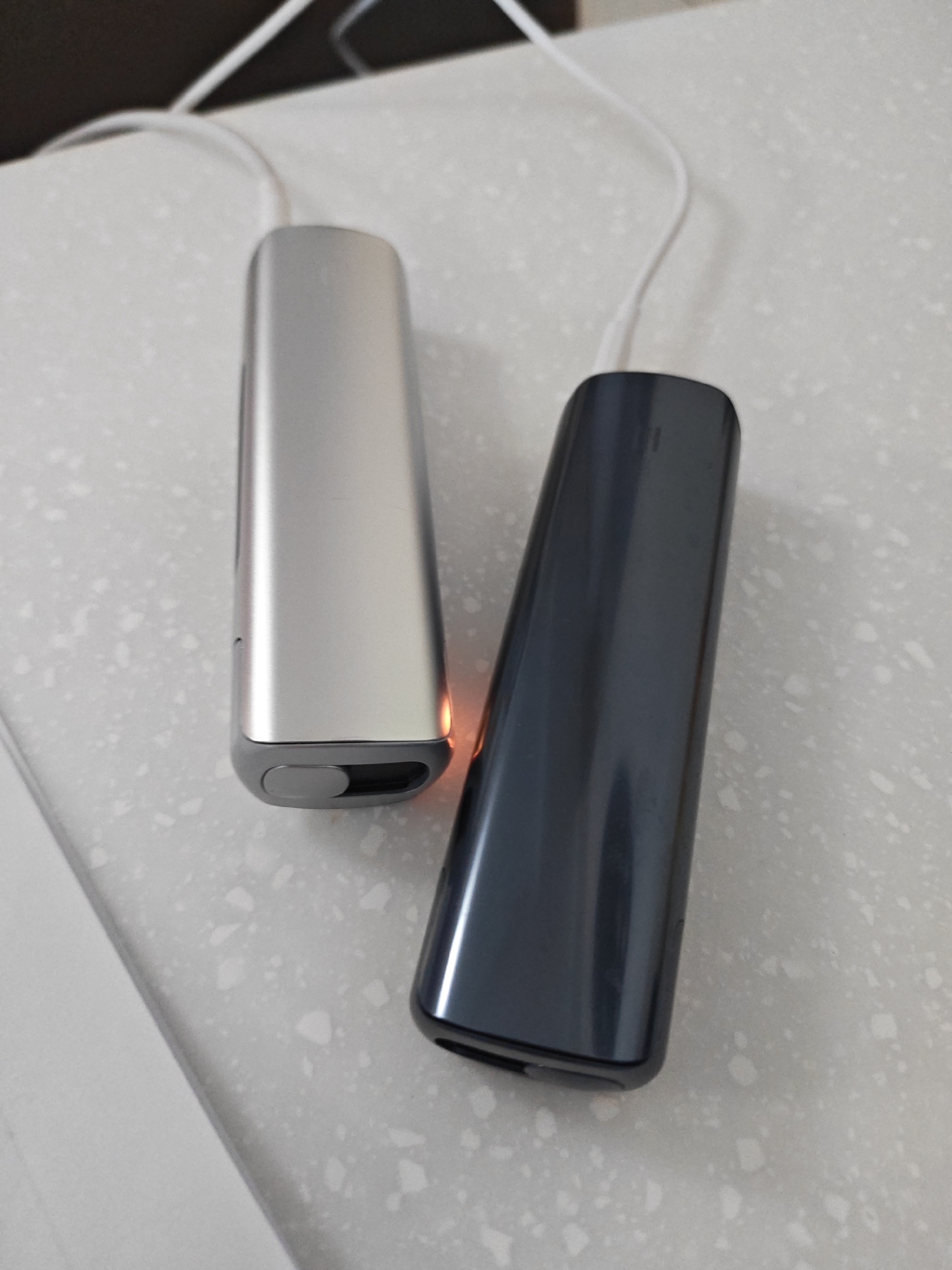 Two electronic cigarette devices, one silver and one dark gray, are placed side-by-side on a speckled white countertop. Both devices have white charging cables connected to them. The silver device is slightly to the left of the dark gray device. The background is blurred and shows a dark surface and a white cable. The scene appears to be indoors, likely in a domestic setting. There is no indication of the location being Vladivostok, Russia.