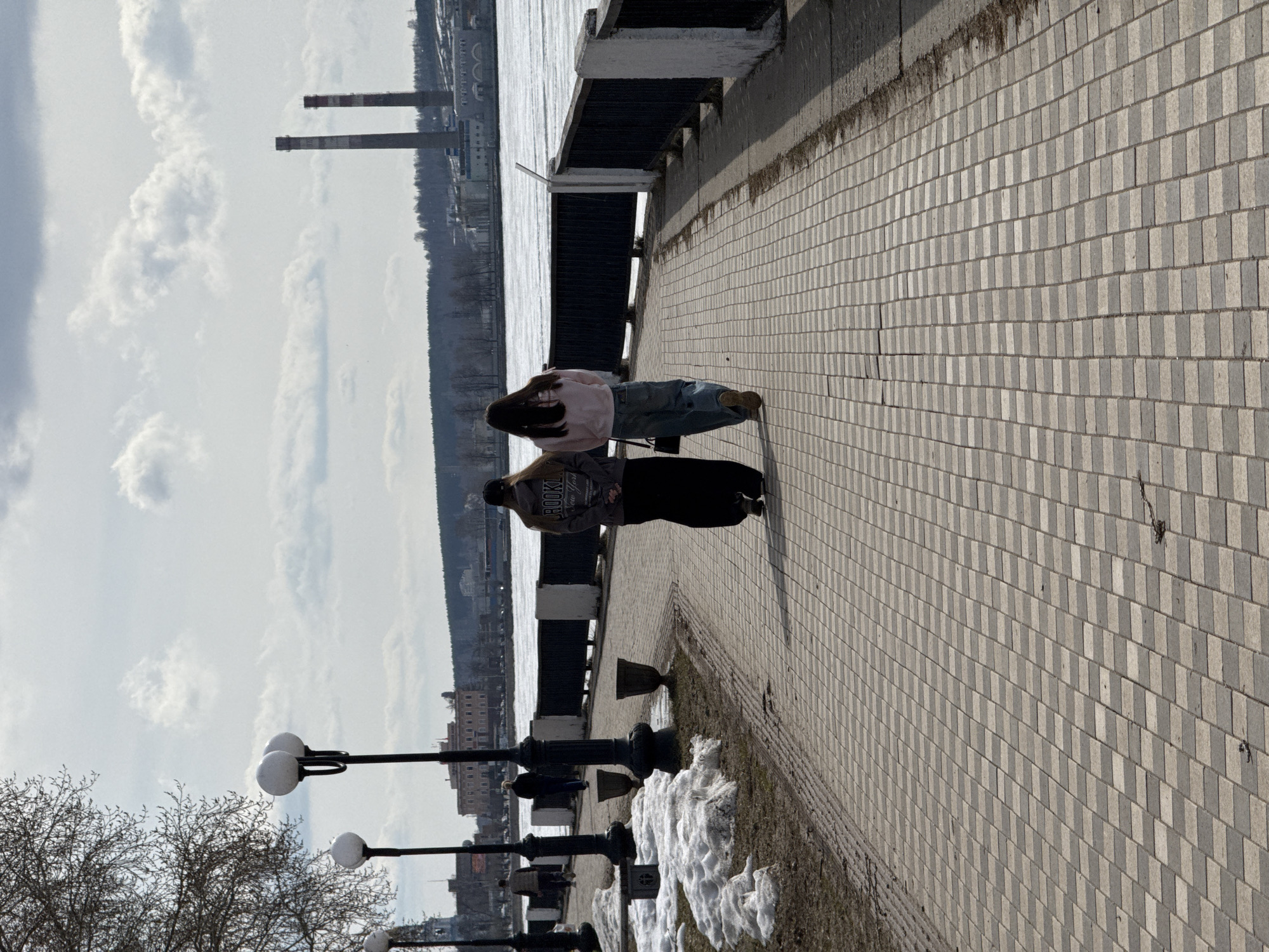 The image shows a waterfront scene in Votkinsk, Russia. Two people are standing on a paved walkway next to a body of water; they appear to be a couple walking their dog. In the background, there are industrial structures, likely chimneys or towers. The walkway is lined with black lampposts with snow at their bases. The weather appears to be overcast.