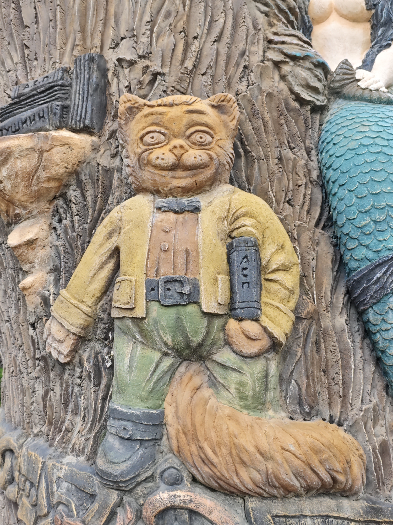 This is a close-up, eye-level shot of a relief sculpture, likely made of concrete or plaster, depicting a whimsical anthropomorphic cat character and a partial mermaid figure. The cat, in the foreground, is rendered in warm, earthy tones of yellow, orange, and brown. It wears a yellow jacket, a brown shirt, green pants, and dark boots. The cat is holding a dark, rectangular object, possibly a book or scroll, in its right hand. The cat's tail, a thick plume of orange, curls around its legs. To the right of the cat, a mermaid is visible. Her upper torso is light-skinned with dark hair, and her lower body is covered in iridescent blue scales, with a dark band around her waist. The background of the sculpture features a textured, bark-like surface, suggesting a tree or natural environment. The relief is located on a building facade in the city of Cherkessk, Russia, specifically on the exterior of the Cherkessk State Historical-Cultural Museum-Reserve named after P. V. Gubin. The sculpture is part of a larger artistic installation that includes figures from Russian folklore and literature.