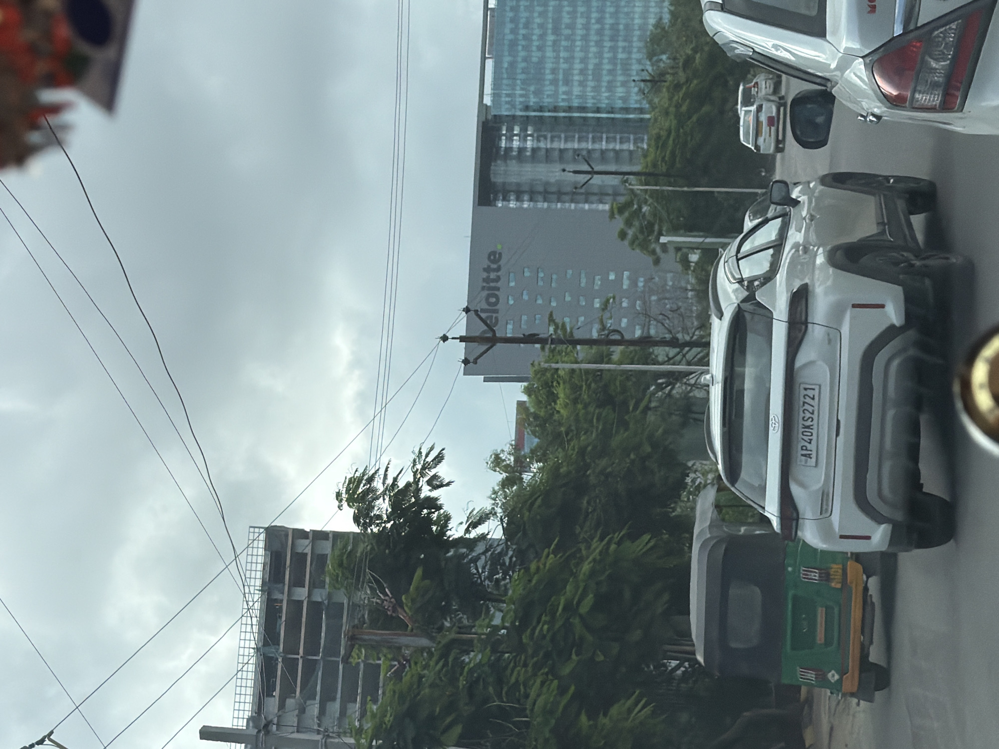 The image captures a street view in Hyderabad, India, on a cloudy day. In the foreground, a white SUV with license plate AP40KS2721 is visible. Behind it, a green auto-rickshaw is also on the road. Further in the background, a modern office building with the "Deloitte" logo is prominent, situated next to another glass-fronted building. The scene also shows a partially constructed building to the left, indicating ongoing development in the area. Power lines crisscross the sky, a common sight in urban Indian landscapes. Trees line the sides of the road, their leaves appearing lush and green. The overall impression is that of a busy urban environment with a mix of old and new infrastructure.