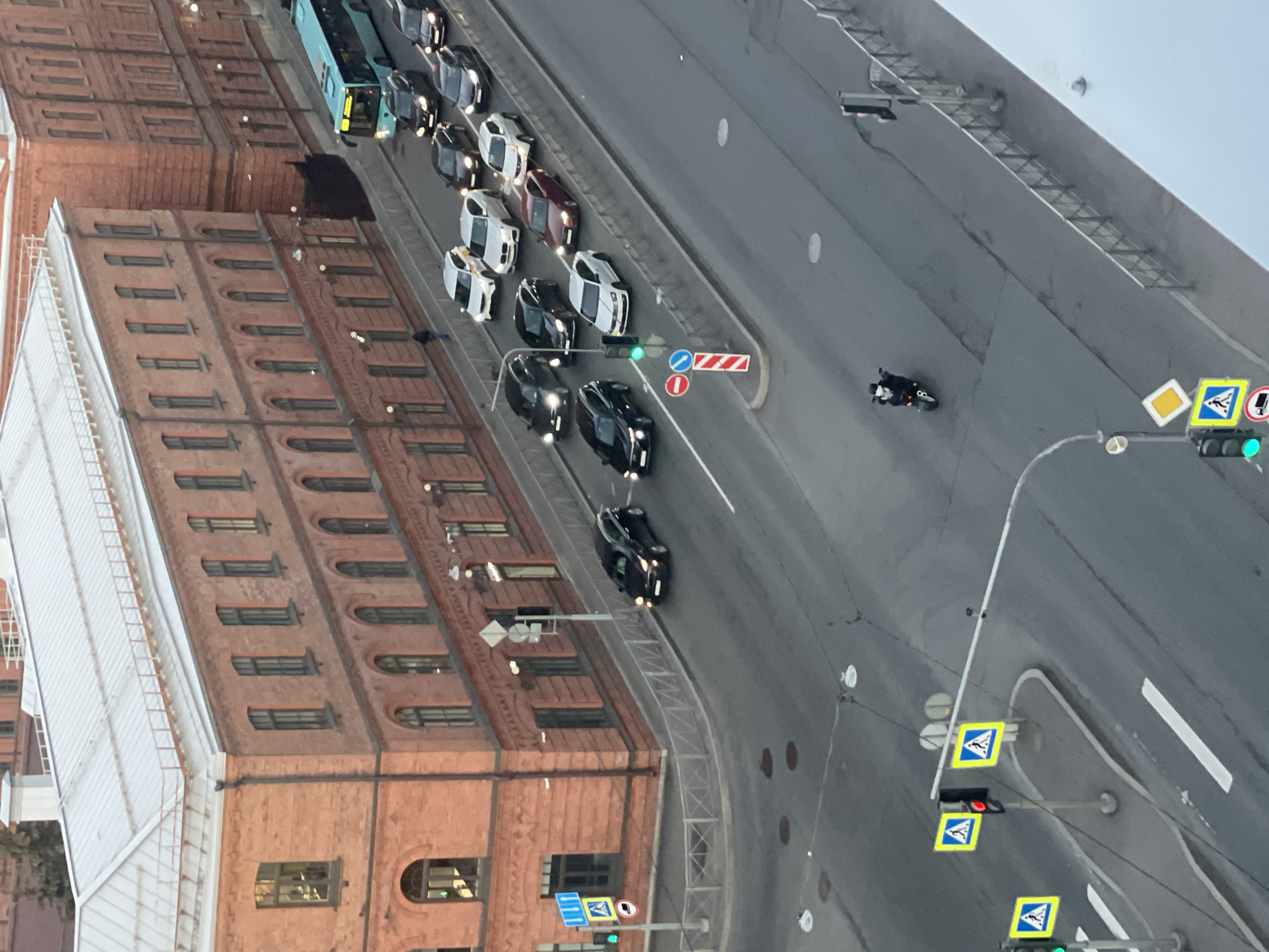 This is a high-angle shot of a street scene in Saint Petersburg, Russia. A red brick building with multiple floors and arched windows is located on the left side of the image. The street is filled with several cars and a bus, primarily dark-colored and white. The traffic appears moderate. A motorcycle is traveling along the street. A series of road signs and traffic lights are visible on the right side of the street. The overall view suggests an urban environment with a mix of historical architecture and modern transportation.