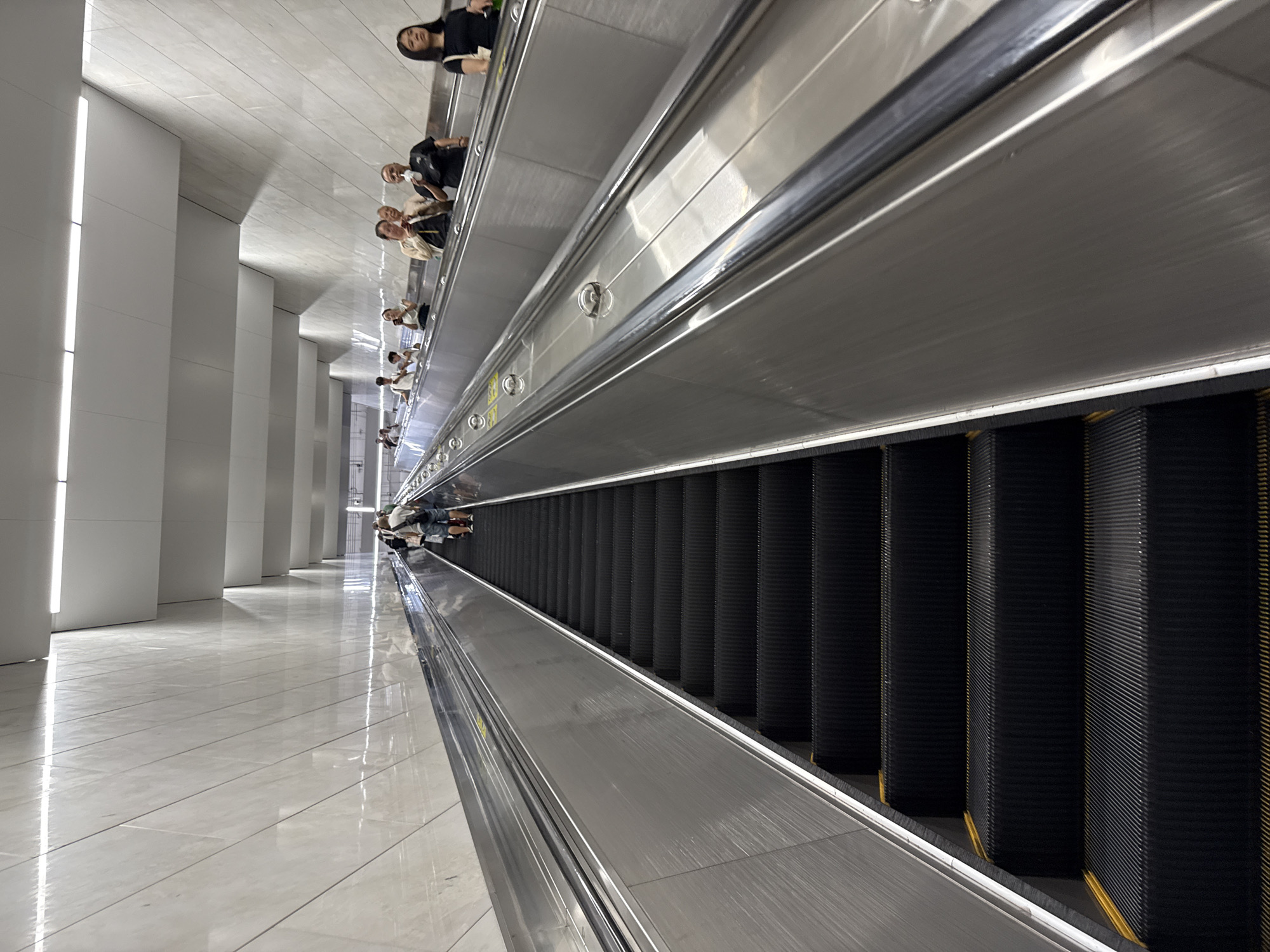 The image captures a perspective looking up a long escalator within the Baiyun District, China. The escalator is made of black, ribbed steps and shiny silver sides. Several people are ascending the escalator, some standing still, while others are in motion. The walls of the station are lined with large, pale marble tiles arranged in diagonal patterns, reflecting the bright overhead lighting. The ceiling features a modern design with layered white panels and recessed lights. The overall atmosphere is clean and contemporary, indicative of a modern transit hub.