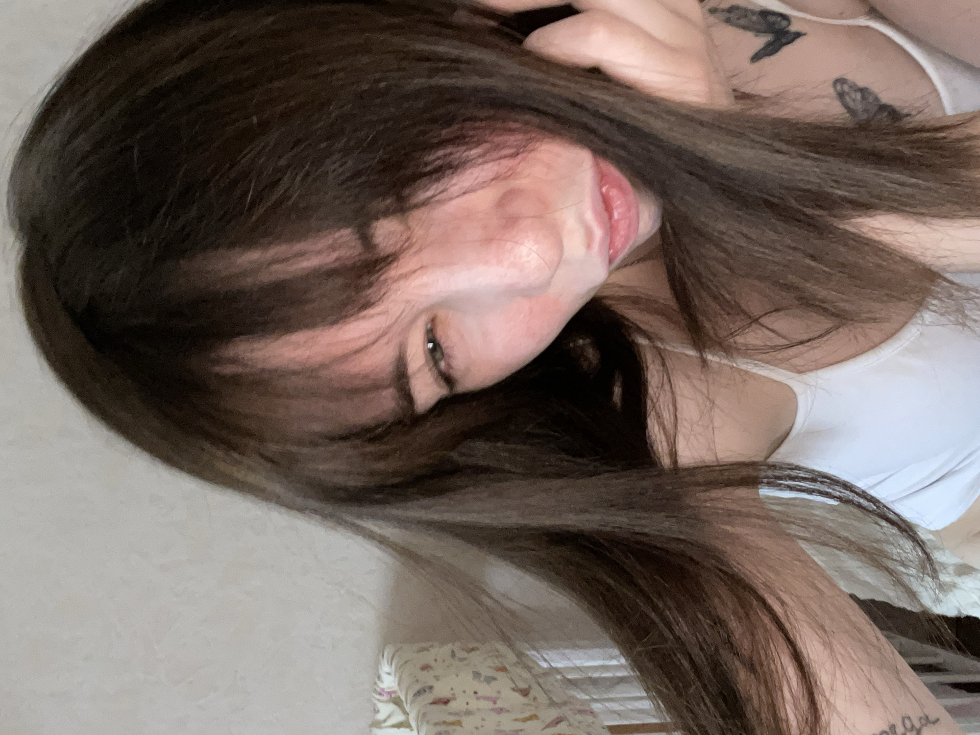 A woman with dark brown hair is the primary subject of the image. She is wearing a white tank top. Two bird tattoos are visible on her upper chest. Part of an arm with text tattooed on it is also visible. The background includes a patterned bedding and a partially visible wall. The location is not discernible based on the image.
