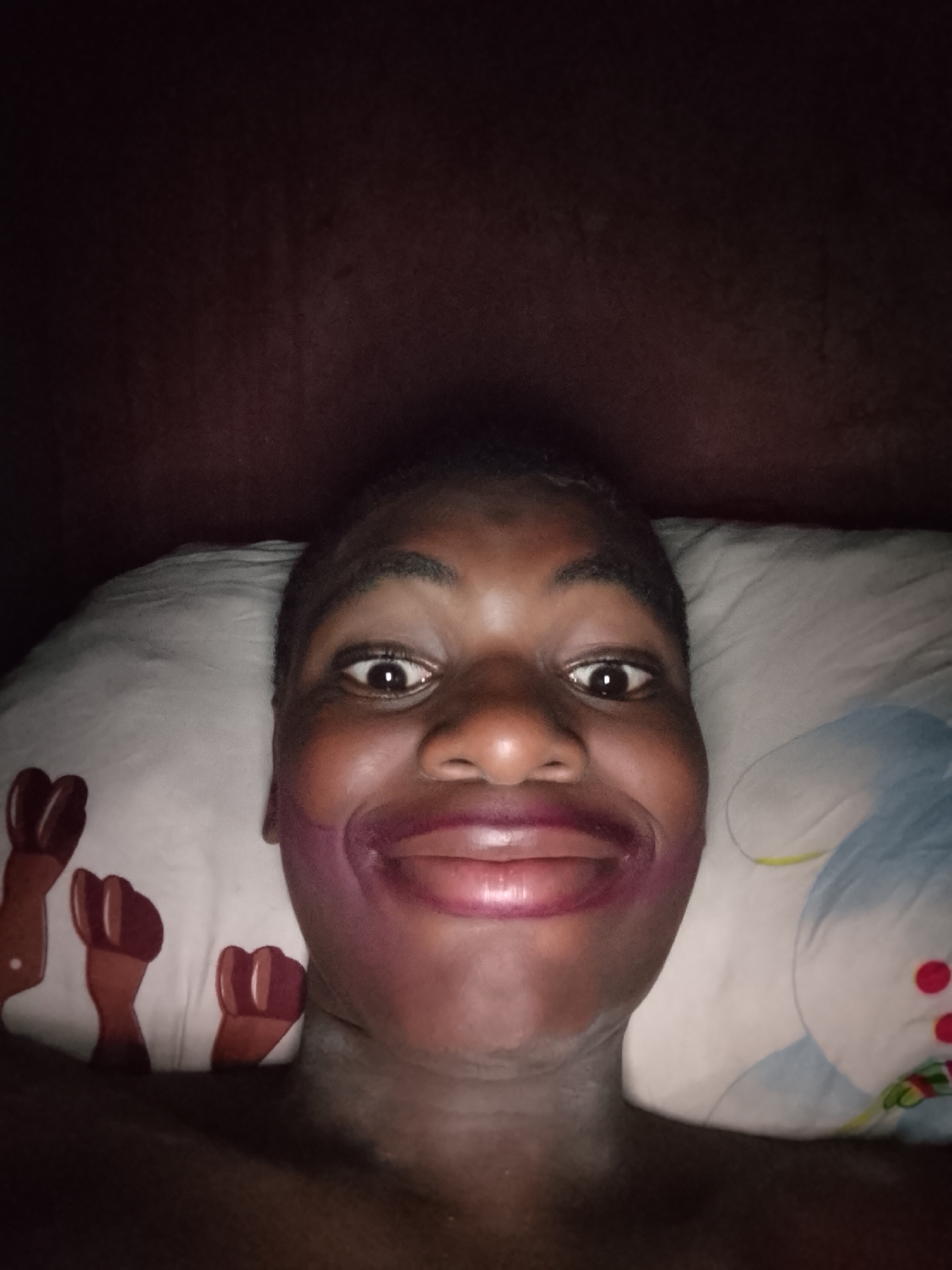 A close-up selfie of a person with dark skin is displayed against a dark, blurry background. The person's face is the primary focus, with eyes wide open and a broad smile. The lips are prominently emphasized with dark purple lipstick that extends beyond the natural lip line. The person is lying on a pillow with a white and light blue design, which features red dots and brown figures resembling clenched fists. The setting is indoors and poorly lit.