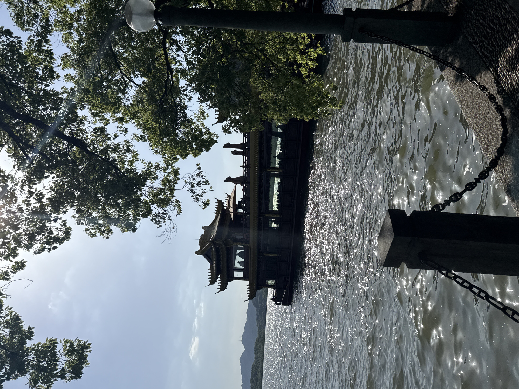 A daytime, low-angle shot depicts a building in the water, potentially in the Xihu District of China. The image includes the building, water with sunlight glinting off it, trees, a street light, and a wall with a chain. The building is a multi-tiered structure with a traditional Chinese architectural style. The water reflects sunlight, creating bright spots on the surface. Trees are visible on the left side of the image, and a street light stands above the water. A wall runs along the water's edge, with a chain hanging from it. Mountains are visible in the background. The sky is pale blue with some clouds.