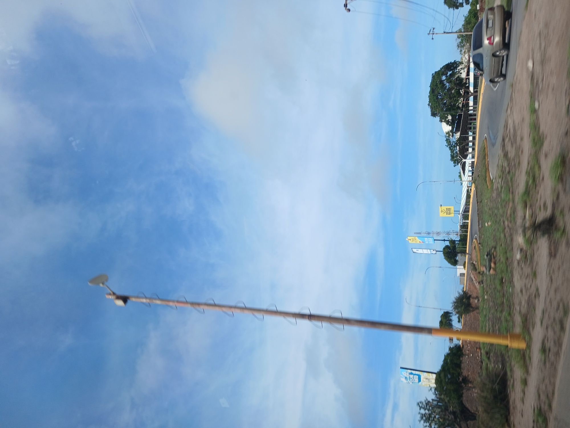 The image depicts a roadside scene under a blue sky with scattered clouds. A tall, weathered utility pole with rungs is prominently featured on the left side of the frame, its top extending towards the sky. In the background, a light-colored sedan drives along a curving road. To the right of the road, a cluster of trees and low-lying vegetation are visible, bordering a building with a light-colored, curved roof. Several billboards with text and graphics are positioned along the road. The overall environment suggests a location in a semi-urban or suburban area, likely in El Valle del Espíritu Santo, Venezuela, given the context. The scene is captured from a moving vehicle, as indicated by the slight motion blur.