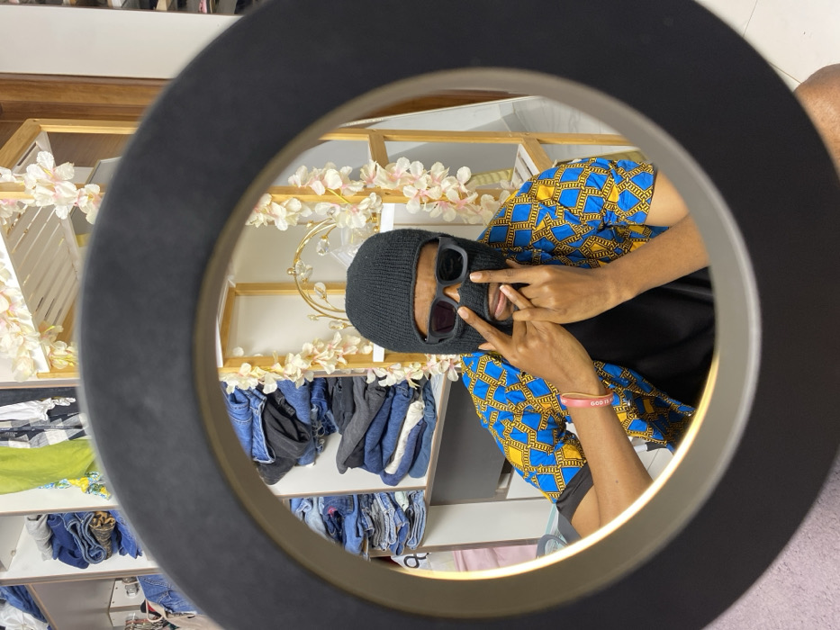 The image depicts an individual within an indoor setting, likely a dressing room or closet in the city of Tema, Ghana. The scene is framed through a prominent black circular object, possibly a ring light, with an illuminated inner edge. The central figure wears a black knitted balaclava covering the entire head and face, with black rectangular sunglasses positioned over the eye openings. They are dressed in a short-sleeved shirt featuring a blue and yellow geometric pattern over a black t-shirt, and a pink wristband is visible on the left wrist. The individual's hands are raised, forming "V" gestures with the index and middle fingers, positioned near the face. The background on the left consists of white shelving units containing stacks of folded clothing, primarily blue denim. To the right, another light-colored wooden shelving unit is adorned with garlands of light-colored artificial flowers, and a gold-colored, ornate decorative object with clear components sits on one shelf.