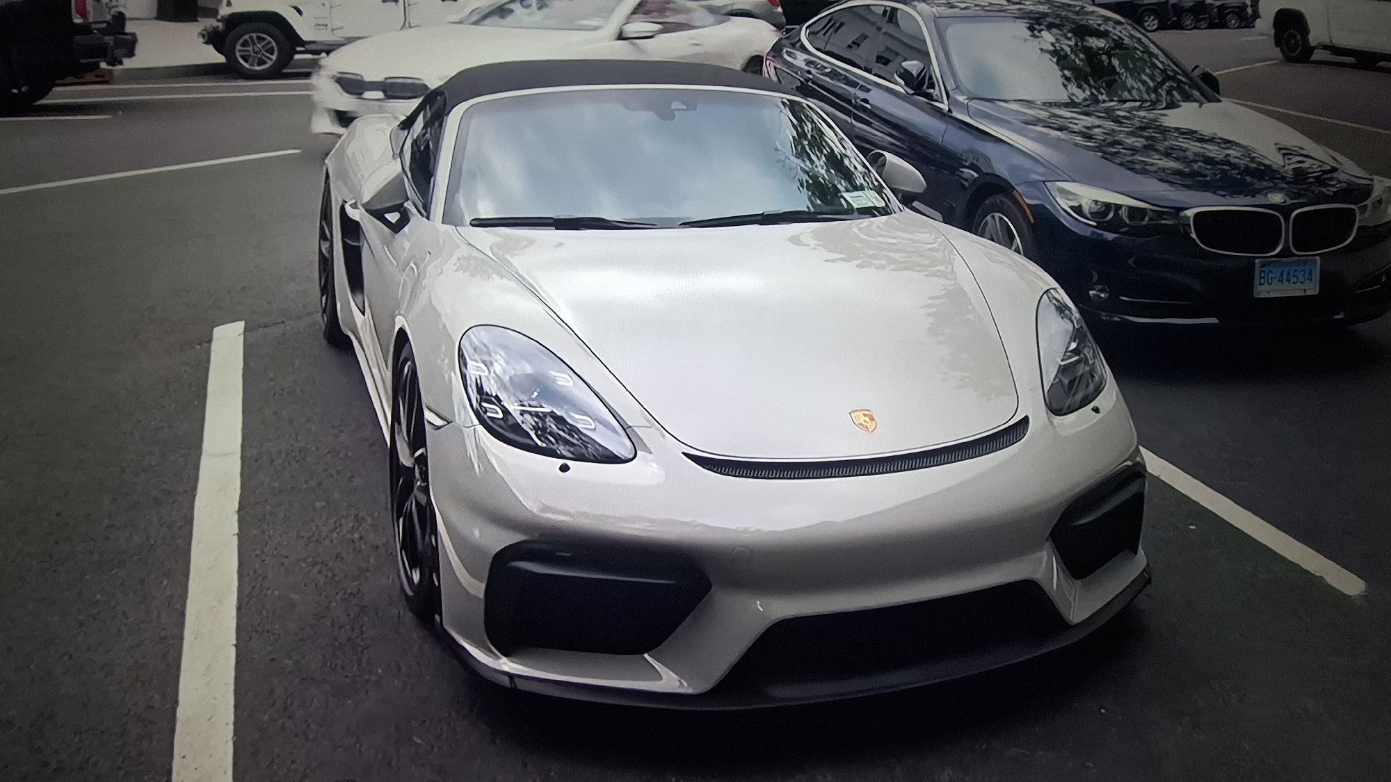 A light grey (or light silver/white) Porsche 718 Boxster Spyder or Cayman GT4 convertible is the central subject, parked at an angle in what appears to be a street or parking lot. The car's front and driver's side are visible, featuring its distinct LED headlights, prominent front air intakes, and the golden Porsche crest on the hood. A black soft top is visible above the windshield.
Behind and to the right of the Porsche, a dark blue BMW 3-series sedan (F30 generation) is parked. Its front license plate is partially visible and reads "BG-44534". Further in the background, a white Jeep Wrangler (JL generation) is partly seen on the left, and another white car, possibly an older generation Toyota Supra, is visible between the Jeep and the BMW. A black pickup truck is also partially visible in the far left background.
The scene takes place outdoors during the daytime, indicated by the bright lighting and reflections of trees and sky on the vehicles' surfaces, suggesting clear or partly cloudy weather. The ground is asphalt with white painted parking or road lines. There are no people visible in the image.