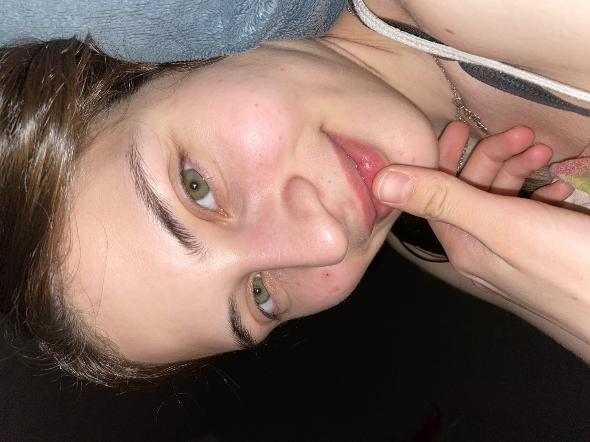 A young woman is shown in a close-up, angled shot. Her face is partially visible with dark brown hair and light green eyes. Her finger is pulling down her lower lip. Part of her neck and chest are visible. A silver chain necklace is around her neck. The background is dark and out of focus. No details are discernable that would suggest this image was taken in Votkinsk, Russia.