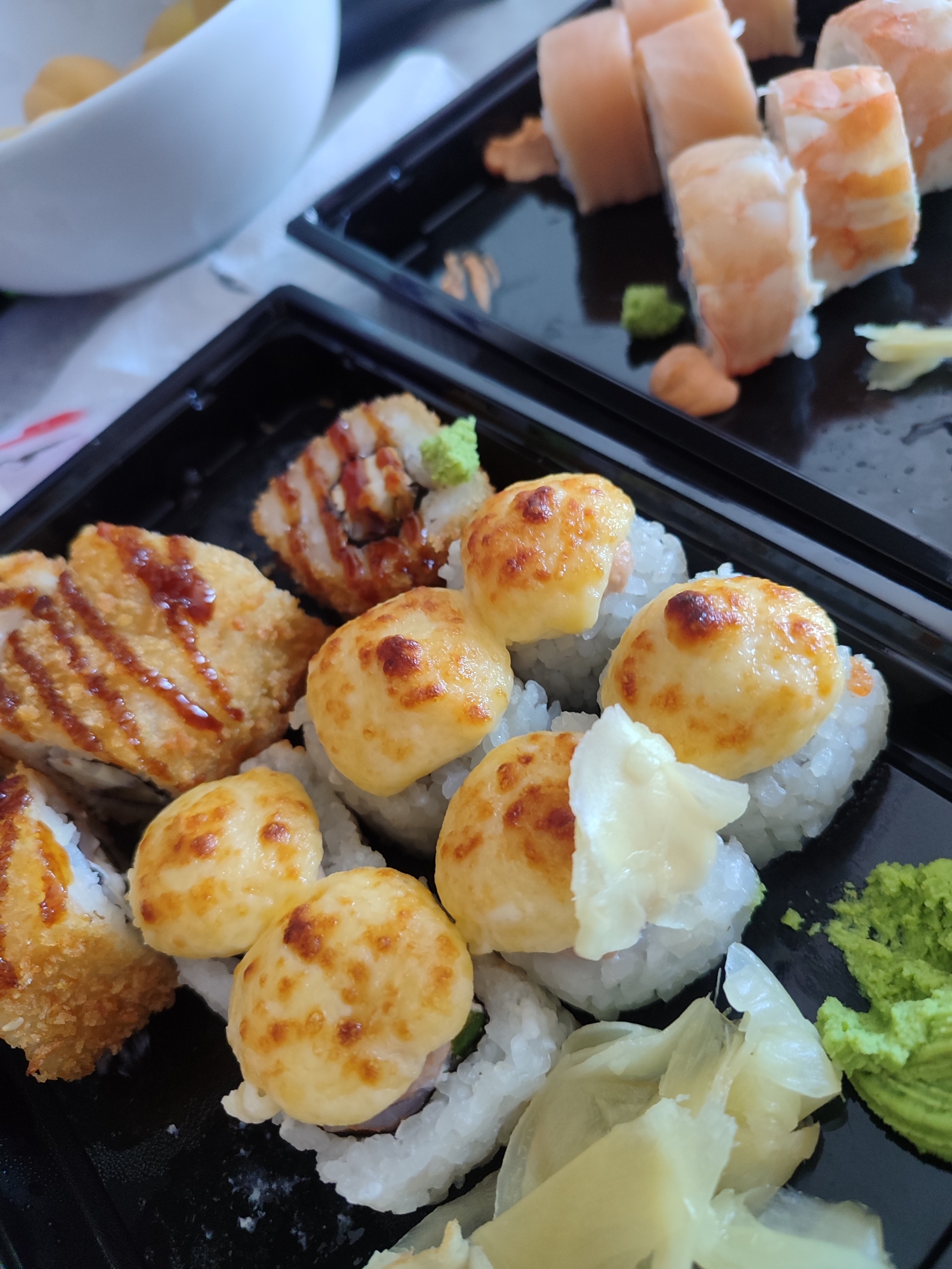 This is a close-up, eye-level shot of two black plastic containers filled with sushi. The container in the foreground is closest to the viewer and is filled with several pieces of sushi. Some of these pieces are topped with a golden-brown, creamy sauce that has been broiled. Other pieces are covered in a crispy, breaded coating and drizzled with a dark sauce. There is also pickled ginger and a dollop of bright green wasabi in this container.
The container in the background, slightly out of focus, contains sushi rolls topped with shrimp. A small amount of wasabi and a creamy sauce are also visible in this container. A white bowl, partially visible in the upper left corner of the image, appears to contain some round, golden-colored food items, possibly dumplings or a similar dish.
The lighting suggests it is daytime, and the setting appears to be a casual meal, likely at home or in a take-out setting. No people are visible in the image. The overall impression is a variety of sushi dishes ready to be eaten. There is no visible text in the image. The location context of Sochi, Russia, does not provide any direct visual cues in the image itself.