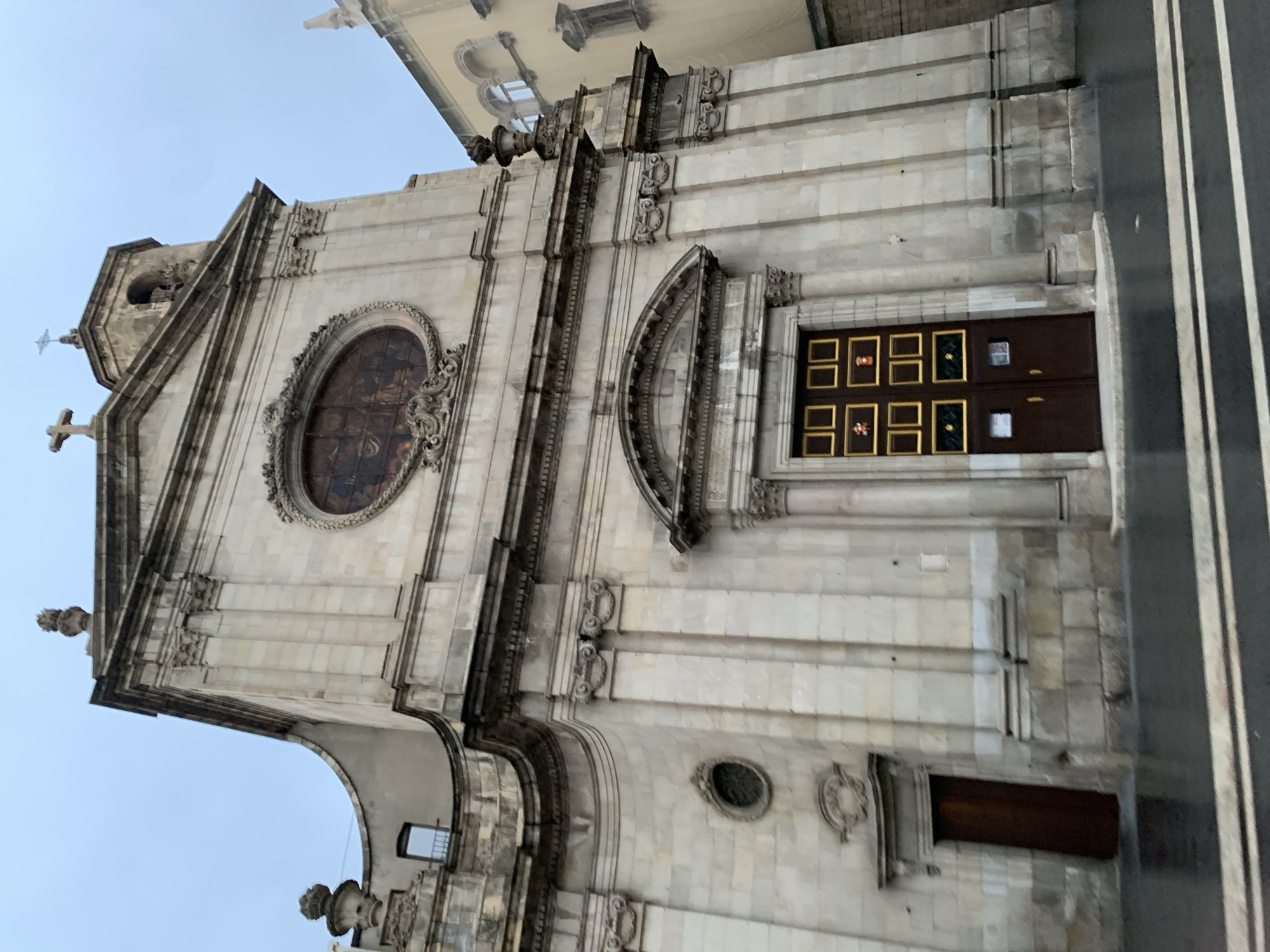 The image shows the facade of a building, likely a church, with notable architectural details. The building is constructed from light-colored stone and features a triangular pediment at the top, adorned with crosses. An oval window with an ornate frame is visible on the upper part of the facade. The main entrance, comprised of wooden doors decorated with gold frames, is situated on the right side of the building. The overall style suggests a historical, possibly Baroque or Neoclassical, architectural design. The building is situated in an urban environment, with a street or sidewalk visible on the right edge of the image. While there is no explicit indication of the location being Barcelona, Spain, the architectural style is reminiscent of buildings found in Barcelona.