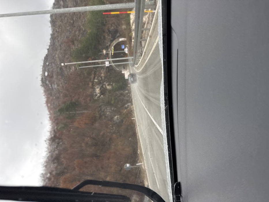 The scene depicts a road in Strugari, Montenegro, viewed from inside a vehicle through a wet windshield, indicating precipitation. The dashboard occupies the lower portion of the frame, with a windshield wiper blade visible on the left. A two-lane asphalt road, wet with water, extends into the mid-ground, curving slightly rightward. White lane markings are discernible. Ahead, a dark-colored sedan travels away from the viewer, its rear visible, generating road spray. The vehicle appears to be approaching a concrete tunnel entrance positioned slightly right of center. The tunnel displays internal lighting and external signage or lights. The surrounding environment consists of steep, hilly terrain covered in a mix of sparse, bare deciduous trees and some evergreen foliage, suggesting a colder season. The sky is overcast and uniformly gray. Road infrastructure includes a metal guardrail system on the right side of the road, multiple utility poles or streetlights, a circular reflective road sign on the left, and a red and yellow striped road marker pole on the far right. No individuals are visible.