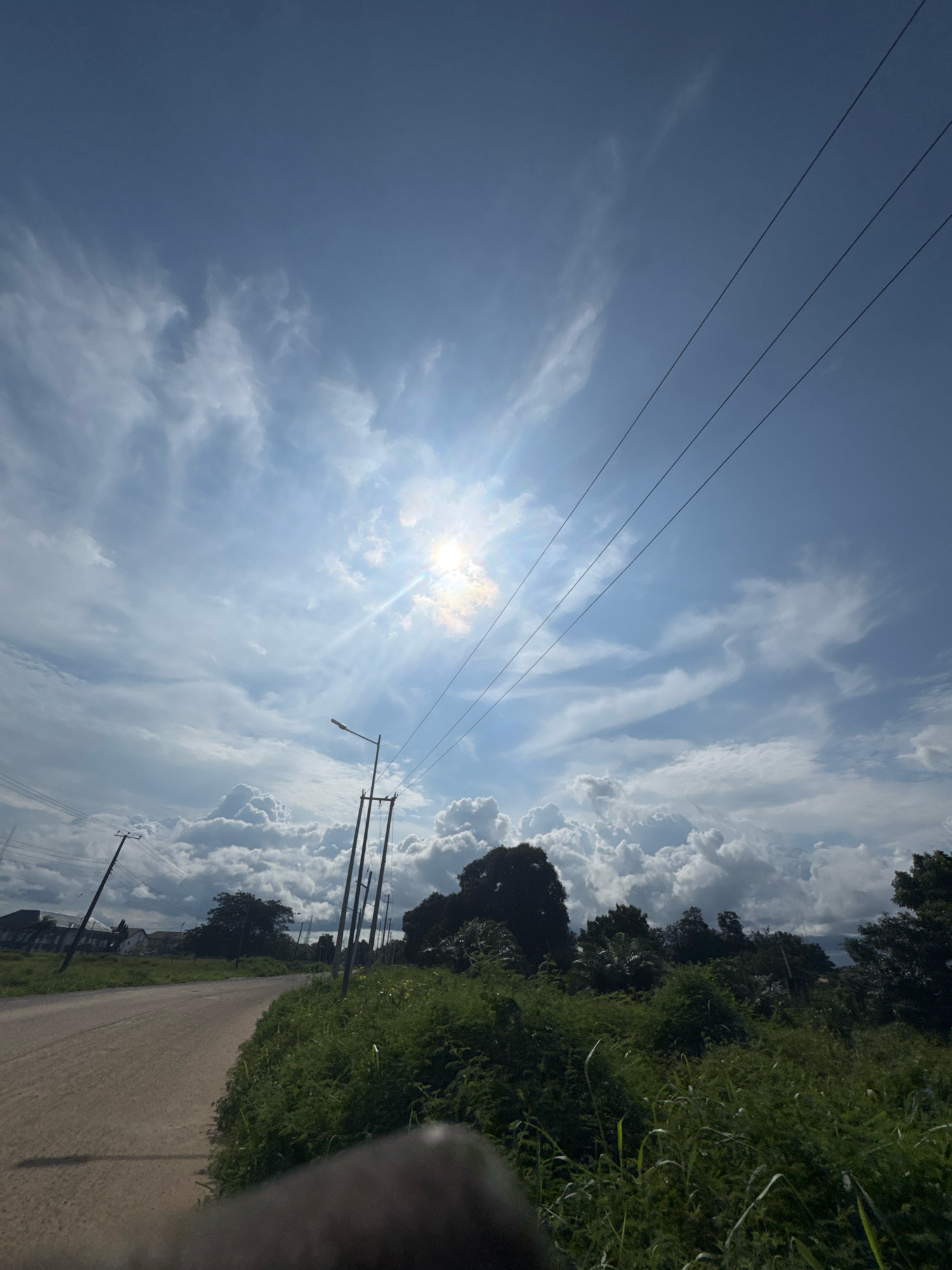 This is a wide-angle, low-angle outdoor photograph taken during the daytime in Ihiagwa, Nigeria. The scene depicts a dirt road curving to the left, bordered by lush green vegetation on the right and a few buildings in the distance on the left. Power lines stretch diagonally across the sky, with a bright sun shining through dramatic, scattered white and grey clouds. Several utility poles with wires are visible along the side of the road. The foreground shows a blurry, dark object, possibly the edge of a camera or other equipment. The overall impression is one of a rural or semi-rural area under a dynamic sky.