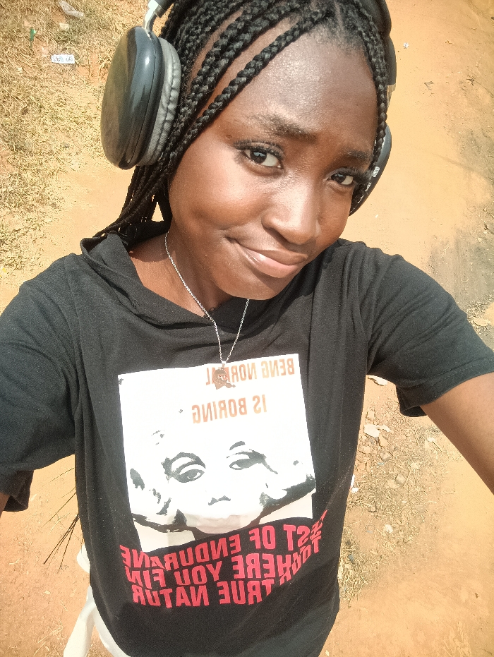 A young Black female is captured in a waist-up selfie, standing outdoors in Oye, Nigeria. She wears black over-ear headphones, a silver-colored chain necklace, and a black short-sleeved hooded t-shirt. The t-shirt features a large white graphic print with a mirrored inscription at the top reading "BEING NORMAL IS BORING," below which is a stylized illustration of a face, and partially visible mirrored text at the bottom. She has dark braided hair, dark eyes, and a slight smile, looking directly into the camera. The background consists of reddish-brown earth with sparse, dry-looking vegetation. A small white object is partially visible in the lower-left foreground.