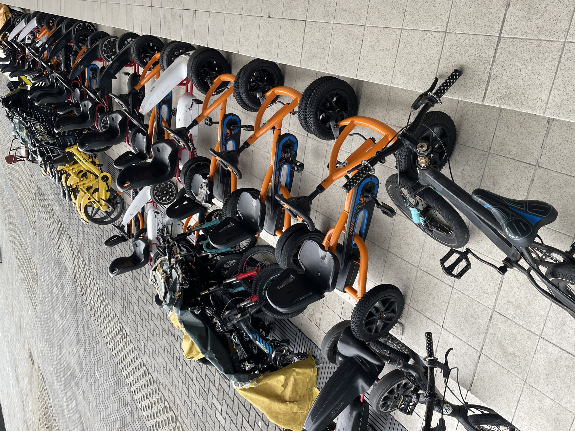 The media file displays a crowded arrangement of various children's vehicles, predominantly tricycles, pedal go-karts, and bicycles, lined up against a light-tiled wall. The scene is viewed from a slightly elevated angle, looking down upon the vehicles and the textured ground.
The majority of the vehicles are orange and black pedal go-karts or robust tricycles, featuring black seats, large black wheels, and occasional blue accents on their frames. Some of these orange vehicles appear to have numbers, such as '22', on their front. Interspersed among them are several yellow balance bikes or small scooters, and conventional children's bicycles in black and light blue. One black bicycle has "BIKE SADDLE" printed on its seat, and another light blue bike shows a partial "RACING" text on its frame. The handlebar grips of one bike are black with small white star patterns.
The vehicles are tightly packed, suggesting a storage area, rental fleet, or a play zone. No people are visible. The ground consists of gray paving, with sections of herringbone pattern and possibly other textured designs. The background wall is composed of light-colored, possibly off-white or very light gray, rectangular tiles. The lighting suggests daytime, possibly overcast or indoors with even illumination, as no strong shadows are cast.