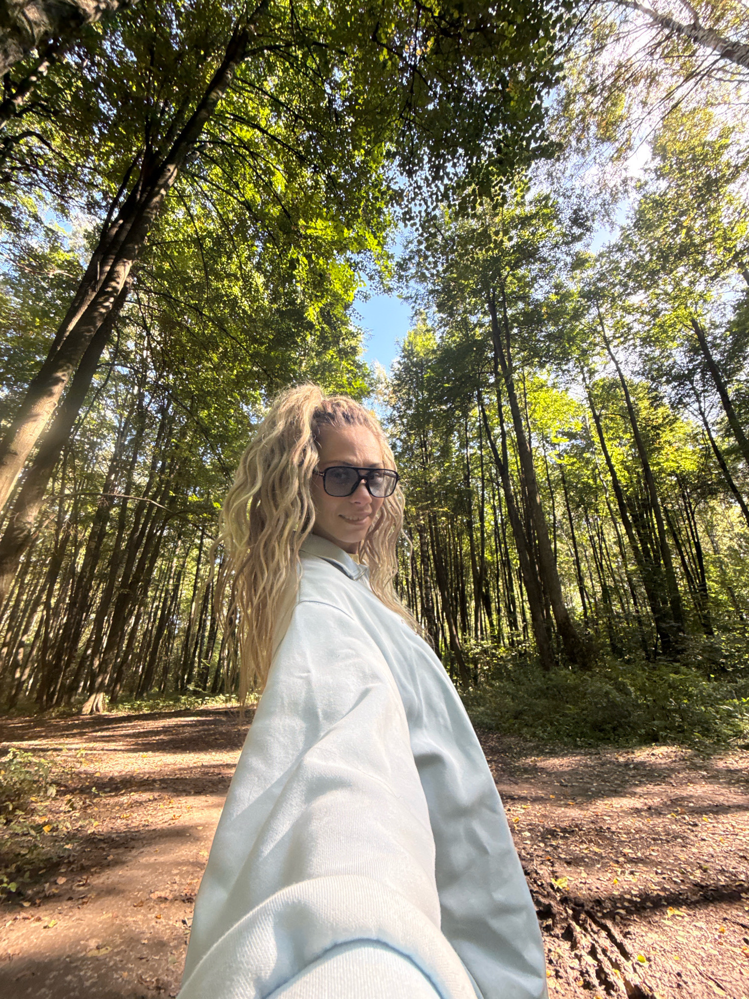 A woman with long blonde curly hair and sunglasses stands in a sun-dappled forest. She is wearing a light blue long-sleeved shirt. The path behind her is covered in fallen leaves. The image is taken from a low angle, looking up towards the canopy of trees. The location is identified as being in the city of Dumino, Russia.